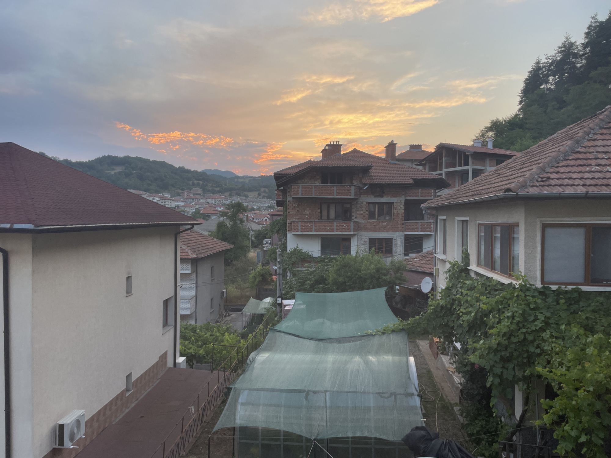 The image depicts a view of rooftops and buildings in the city of Zlatograd, Bulgaria, during what appears to be sunset. The sky is filled with colorful clouds, ranging from pale orange to gray. Several residential buildings with tiled roofs are visible, some appearing to be under construction. In the foreground, a large green mesh covers what looks like a greenhouse or a protected garden area. Trees and vegetation are present on the surrounding hillsides and around the buildings. The overall scene suggests a residential area on the outskirts of the city, with a backdrop of natural landscape.