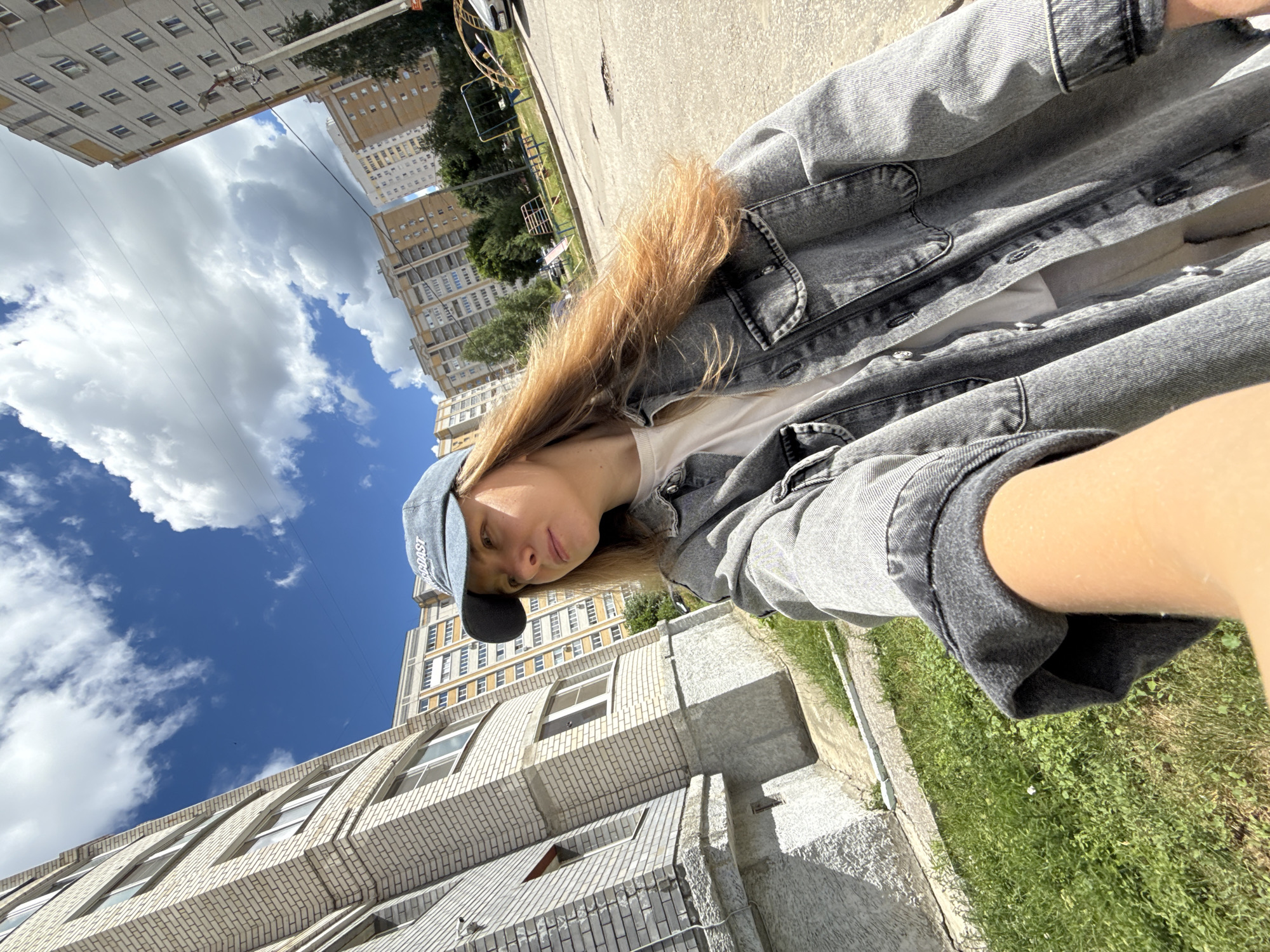 A young woman wearing a grey denim jacket and a blue baseball cap is standing on a paved area in front of a multi-story apartment building. She is looking to her left, away from the camera. The building behind her is made of light-colored brick. In the background, there are other apartment buildings, trees, and a playground with a slide and a swing set. The sky is blue with some white clouds. The ground is cracked and uneven in some places.