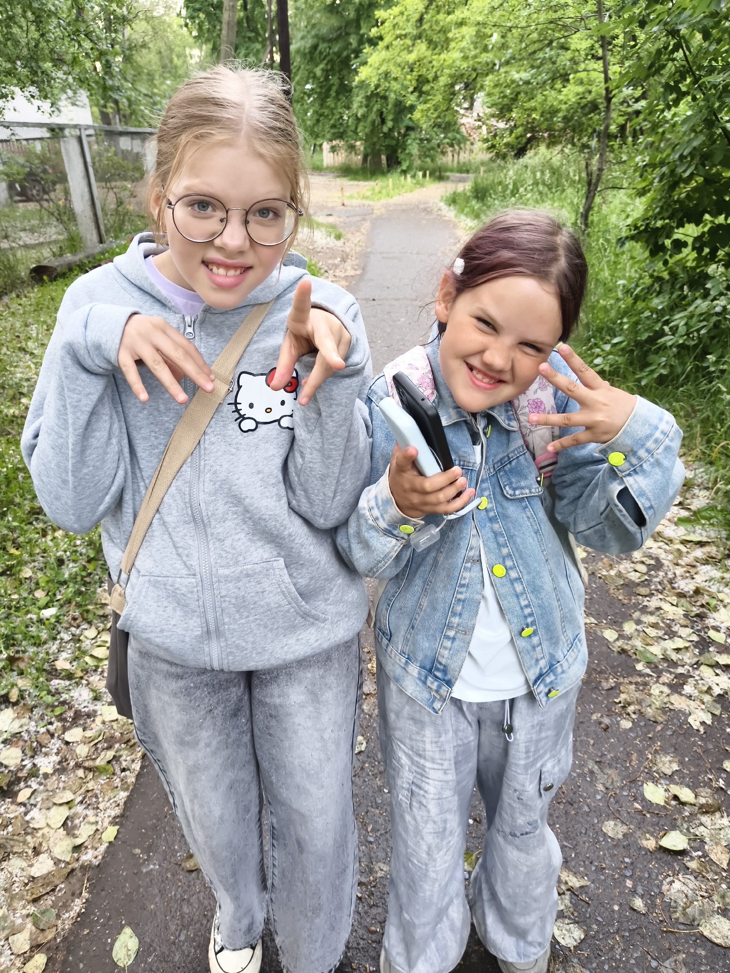 Two young girls, one with blond hair in a grey hoodie and glasses, and the other with brown hair in a denim jacket, stand on a dirt path lined with green trees and fallen leaves. The girl on the left has her hands up, forming claw-like shapes with her fingers, and is smiling at the camera. She wears a grey hoodie with a Hello Kitty graphic on the left breast. The girl on the right holds a black and a light blue object, possibly a phone and a power bank, in her left hand, while her right hand is raised in a peace sign. She is also smiling widely. Both girls are wearing casual clothing, including comfortable-looking pants. The background suggests an outdoor, possibly park-like setting, with a fence visible behind the girl on the left and dense foliage on either side of the path. The ground is wet in places and scattered with leaves, indicating recent rain or damp conditions.