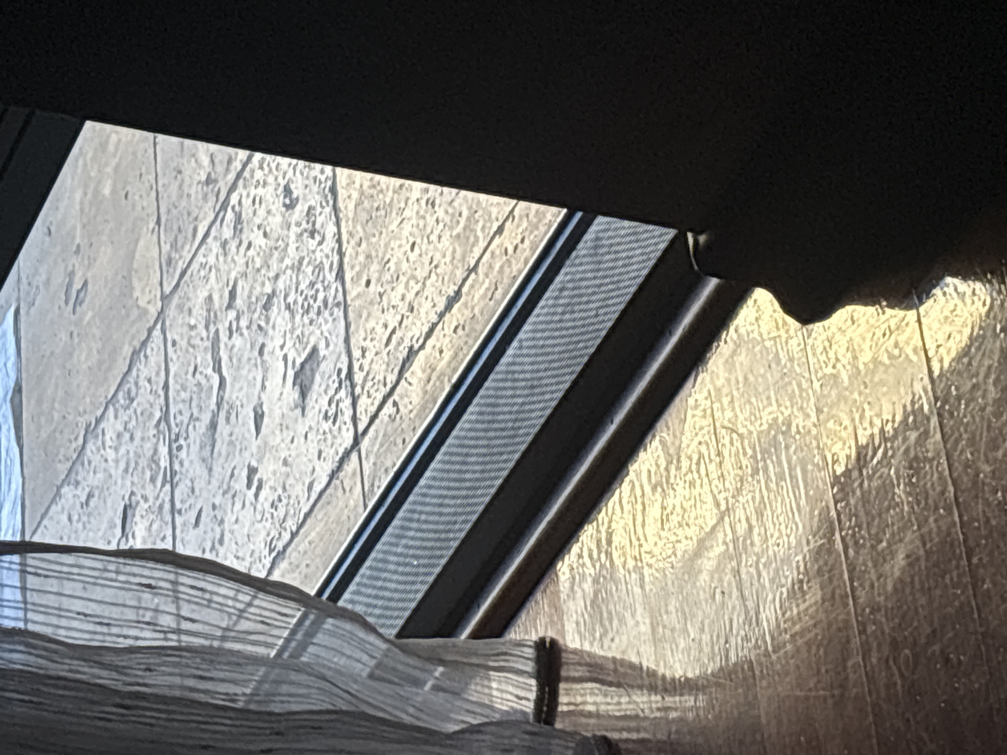 The image shows a partial view through a window in what appears to be an interior space. Outside, the scene depicts light-colored stone walls or pavement, possibly exterior cladding. The light suggests it is either early morning or late afternoon. A section of window frame and screen is visible, along with a portion of fabric or curtain in the foreground. The wooden surface shows light reflection. Budva is not discernible from the image.