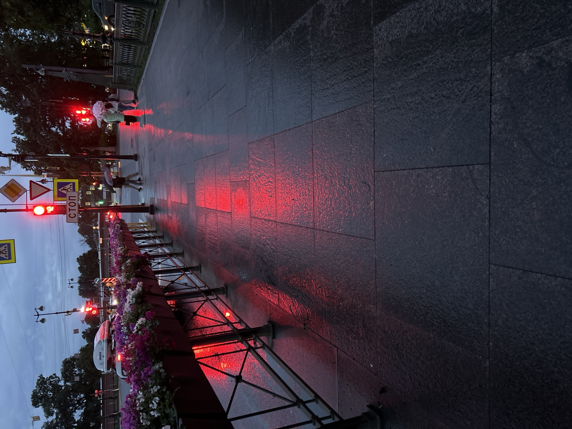 A street scene unfolds at dusk or early morning with a reddish hue cast by streetlights reflecting off the wet pavement. The ground is paved with large, dark gray rectangular stones that are wet, creating a shimmering surface. To the left, a metal railing supports planters filled with blooming flowers, primarily in shades of purple, pink, and white. Red traffic lights are visible in the background, contributing to the overall red illumination of the scene. A white car with a roof-top carrier is parked on the street. Several figures are present, some walking with umbrellas, suggesting it may be raining. One person holds a light pink umbrella, while another holds a dark umbrella. The background features trees and more streetlights, indicating an urban setting. Signs, including a "STOP" sign and traffic signals, are also visible.