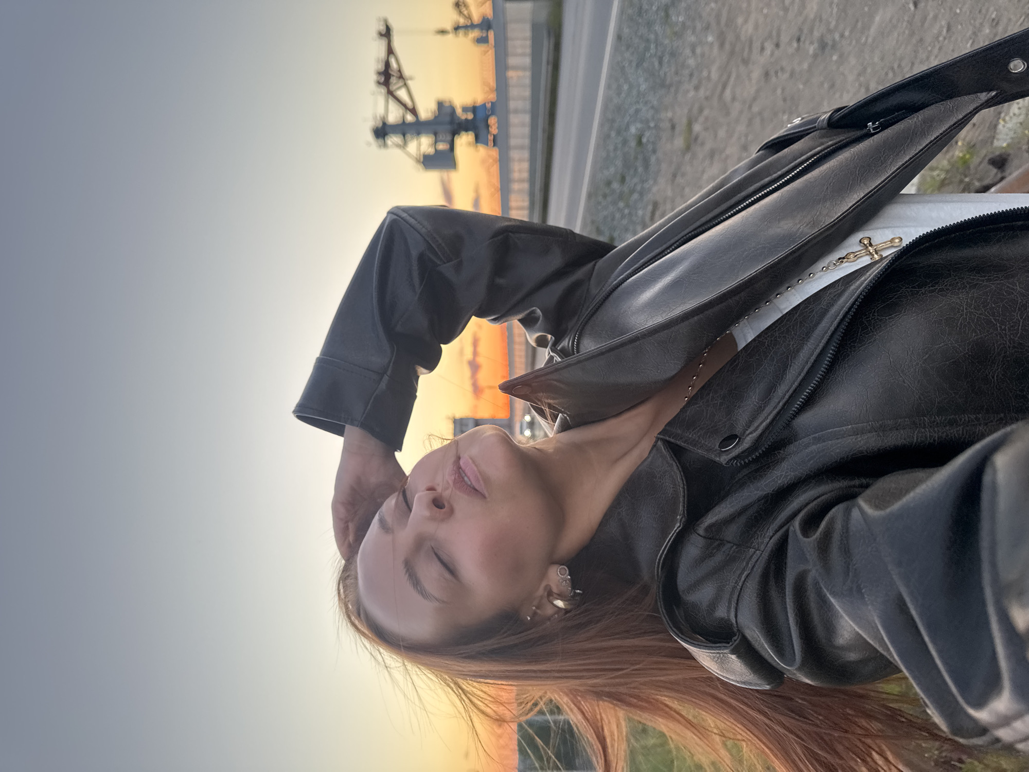 A woman with long reddish-brown hair stands outdoors, her eyes closed and her hand resting on her forehead, against a backdrop of a setting sun and a clear sky. She is wearing a black leather jacket over a white top, adorned with a gold cross necklace. In the background, industrial cranes are visible, along with a road and a grey gravelly surface. The scene appears to be near a port or industrial area, potentially in Chaikovsky, Russia, given the presence of large cranes and the general atmosphere. The lighting suggests it is dusk.