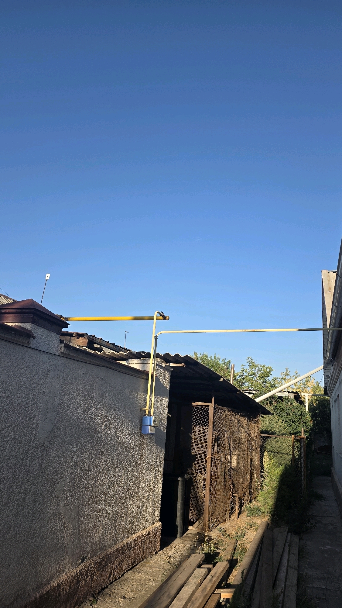 This is a photograph taken from a low angle, looking upwards towards a clear blue sky. The foreground features a narrow, overgrown pathway bordered by wooden planks on the right. To the left is a textured, off-white wall with a dark brown foundation. A yellow gas pipe runs horizontally above the wall and then descends vertically, attached to a blue gas meter.
Behind the wall, a dark, corrugated metal roof extends over a structure with a rusty wire mesh enclosure. Beyond this structure, dense green foliage is visible. To the far right, the side of a building with a white wall and a slanted roof is visible, leading into the distance along the pathway. The lighting suggests it is daytime, with shadows cast on the ground and parts of the structures. The scene appears to be an outdoor area, possibly a backyard or alleyway, in a residential setting. The image does not contain any people or explicit indications of the city of Chuy, Kyrgyzstan, beyond the prompt's assertion.