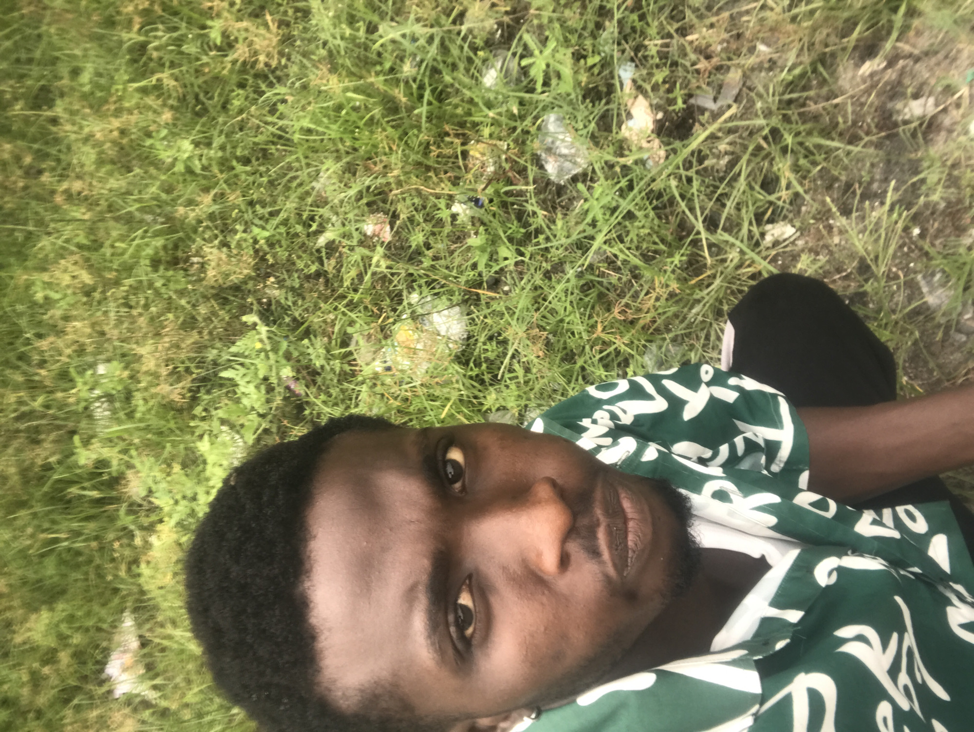A medium shot shows a Black male from the chest up, wearing a green short-sleeved button-down shirt with a white abstract pattern over a white t-shirt. The man has short, dark, curly hair and a short beard. He is looking directly at the camera with a neutral expression. The background consists of overgrown green grass with some scattered debris, suggesting an outdoor environment possibly in a less maintained area. The lighting appears to be natural daylight.