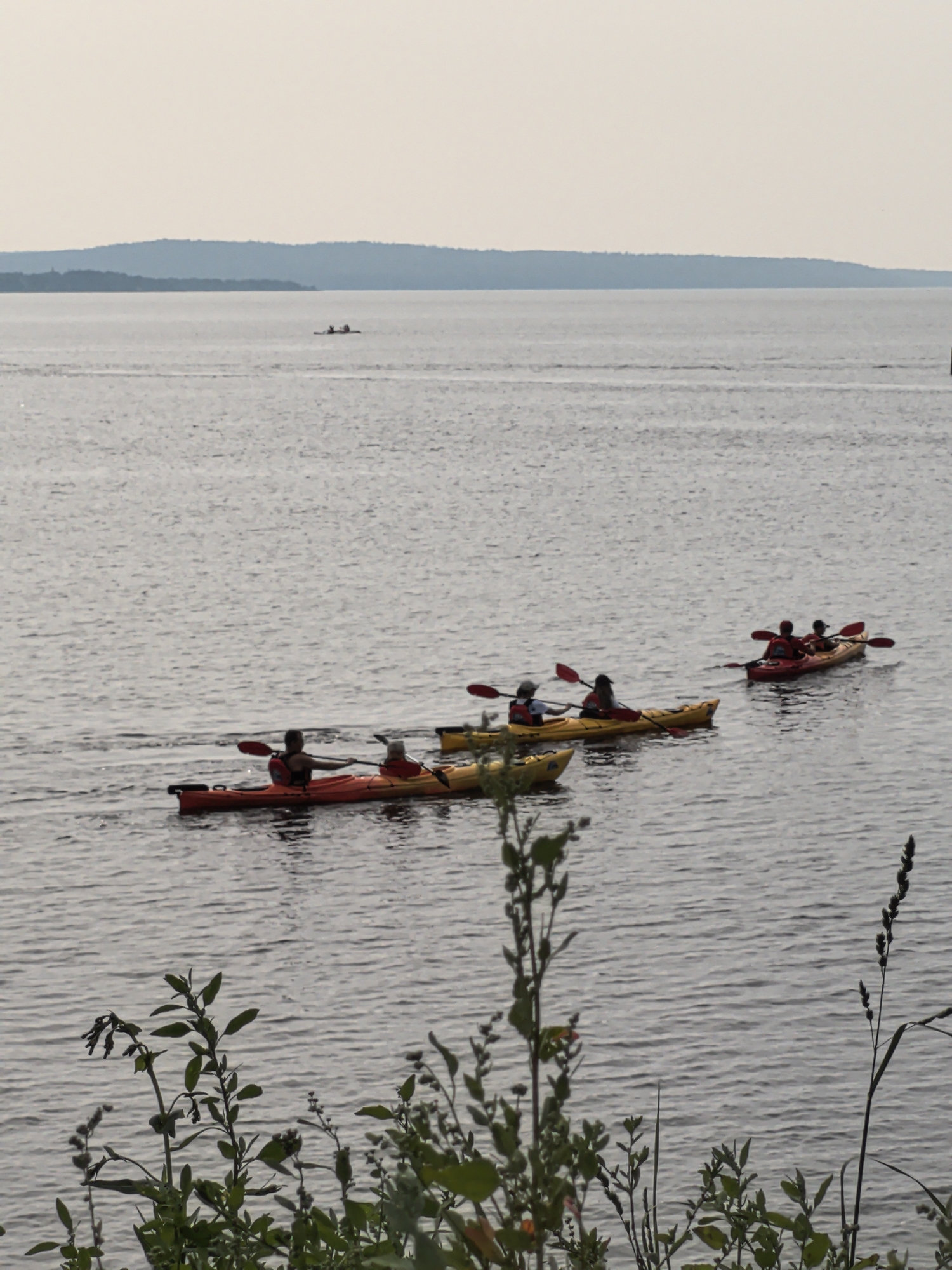 In a serene body of water, four kayaks navigate the calm surface under an overcast sky. The kayaks are positioned in a staggered formation, with the closest one on the left being an orange double kayak carrying two individuals. To its right and slightly behind, a yellow double kayak holds two people, also paddling. Further in the distance, a red double kayak carries two people, and behind that, another red kayak with two occupants is visible. The kayaks are accompanied by their respective paddles. The environment is a vast expanse of water stretching towards a distant, tree-lined horizon. The foreground is occupied by lush green vegetation, with some plants extending into the frame. The scene appears to be a peaceful outdoor activity taking place on a lake or river. There is no information available regarding the specific location in the city of Osa, Russia.