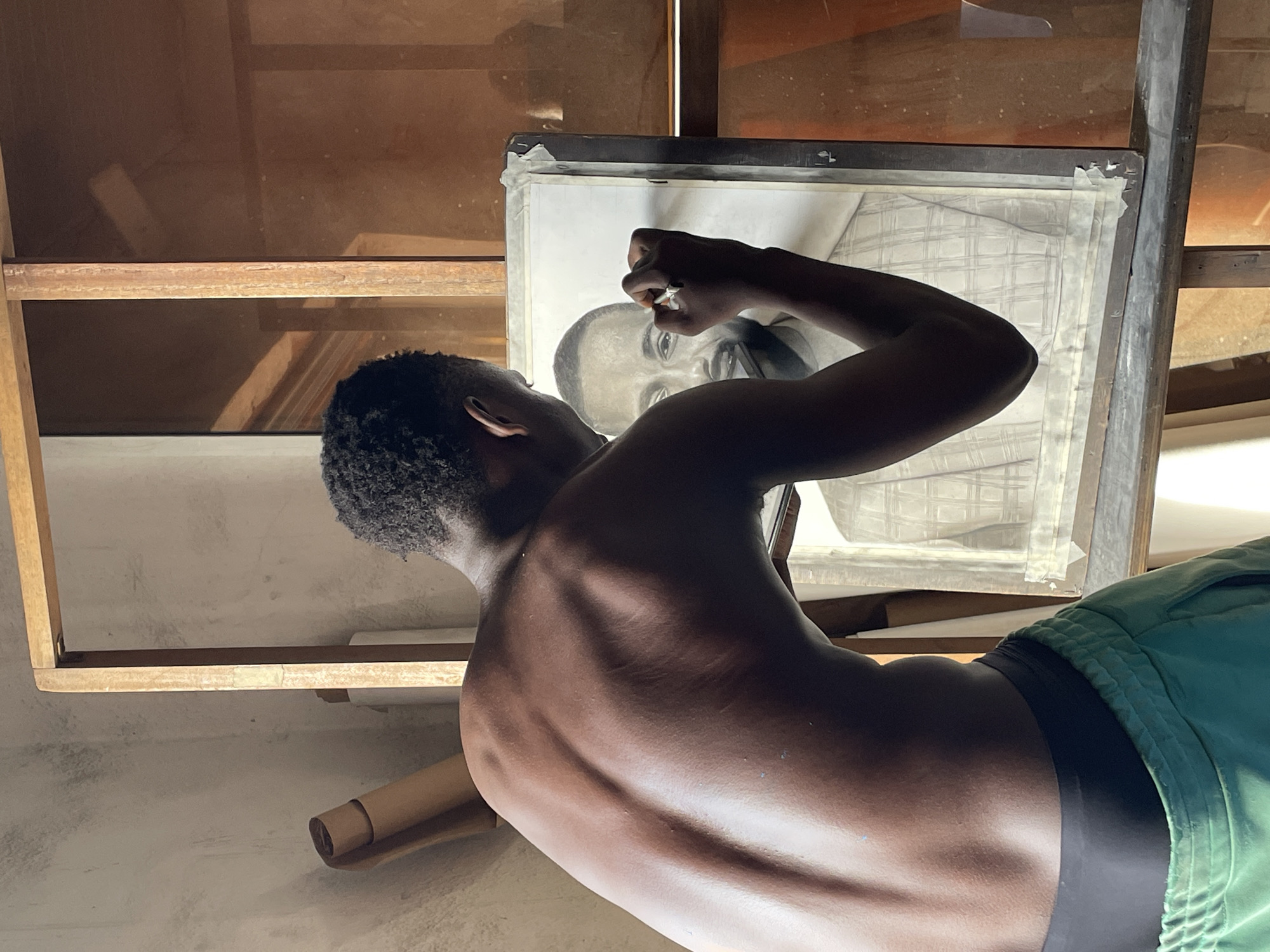 A dark-skinned person is drawing a portrait. The person is unclothed from the waist up, and wears green shorts. The portrait is on a paper taped to a black framed board. The portrait is of a man. The location is inside a room with a wooden frame. The city is Onitsha, Nigeria.