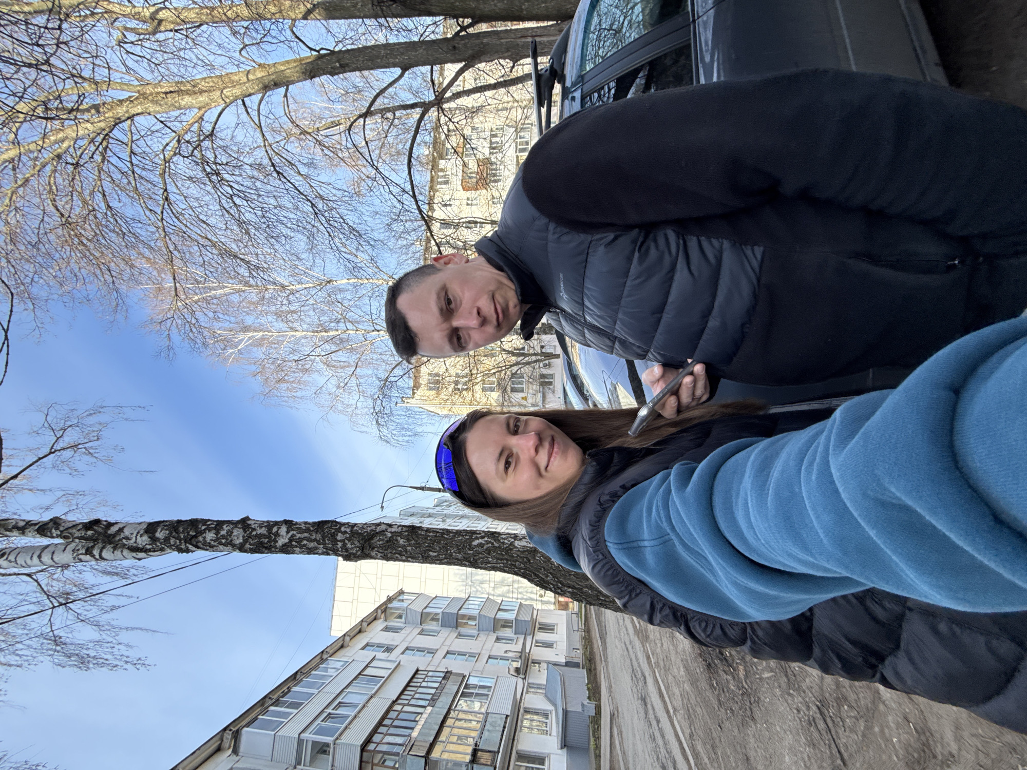 Two individuals, a man and a woman, are captured in a selfie outdoors. The woman, positioned closer to the camera, wears a blue jacket and a blue headband. The man stands behind her, dressed in a black jacket. They are both looking directly at the camera. The man is holding a phone.
Behind the subjects, there is a residential building of approximately 8 floors. Its facade is white with gray window frames. In the background, there are trees with bare branches. The sky is blue with some visible clouds.
The scene appears to be on a sidewalk, next to a parked gray car. Based on the available information and the visual characteristics, the location is likely in the city of Chaikovsky, Russia.