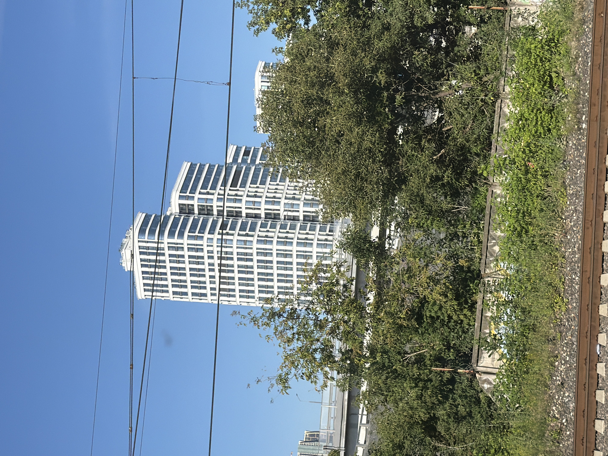 A street-level, outdoor, eye-level shot shows a clear, blue sky with three black electrical wires stretched across the top of the frame. In the background, a modern, white, high-rise building with many windows is visible. In front of this building, there is a green, leafy tree. Below the tree, a concrete wall with graffiti is visible, and in the foreground, a set of train tracks with ballast is present.