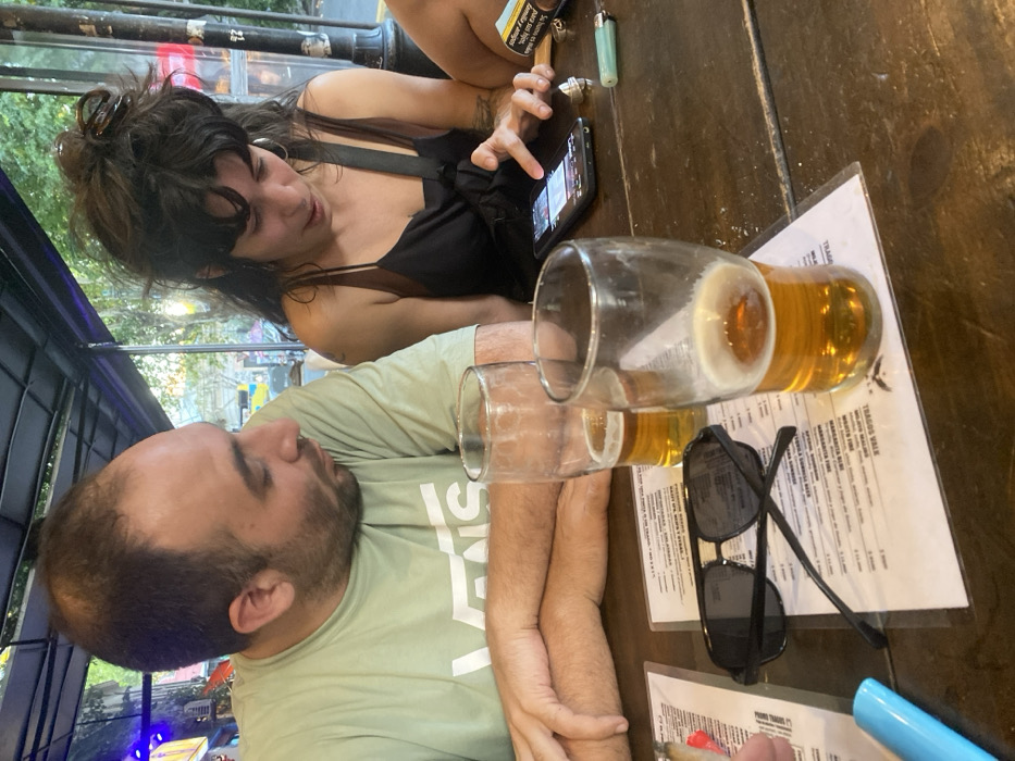 A man and a woman sit at a wooden table. The woman has dark hair, wears a black top, and looks at a phone. The man has a beard and is wearing a green shirt with "VANS" on the sleeve. Two glasses, one mostly empty, the other half full of beer, sit on a menu. Sunglasses and a pen are also on the table. A lighter, a pack of cigarettes, and a second menu can be seen. A partial view of an outdoor setting is visible in the background.