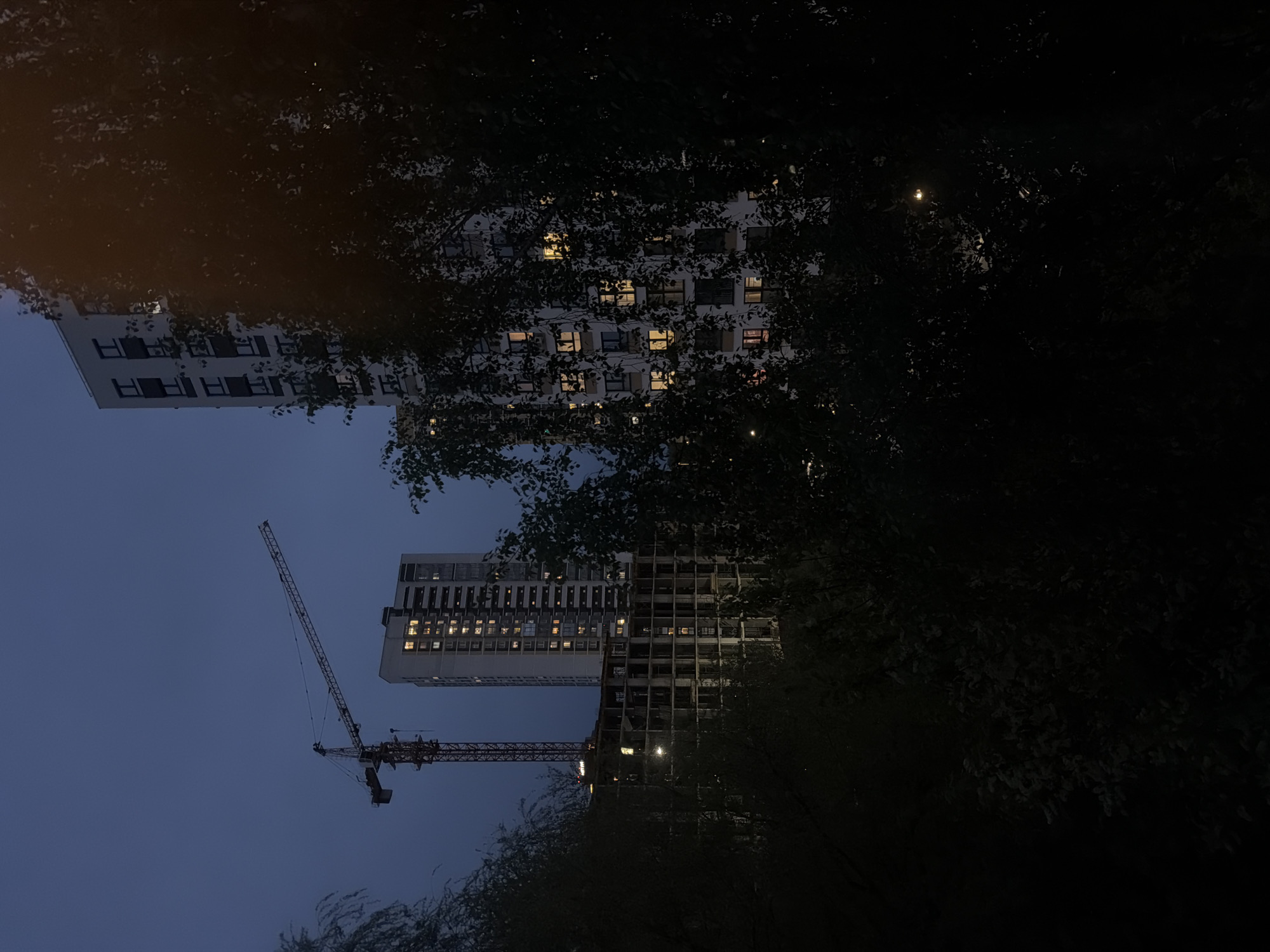 The photograph shows two tall buildings in the city of Izhevsk, Russia, taken at dusk or night. One building appears to be complete, with lit windows indicating occupancy, while the other appears to be under construction. A construction crane is visible near the building under construction. The scene is partially obscured by trees.