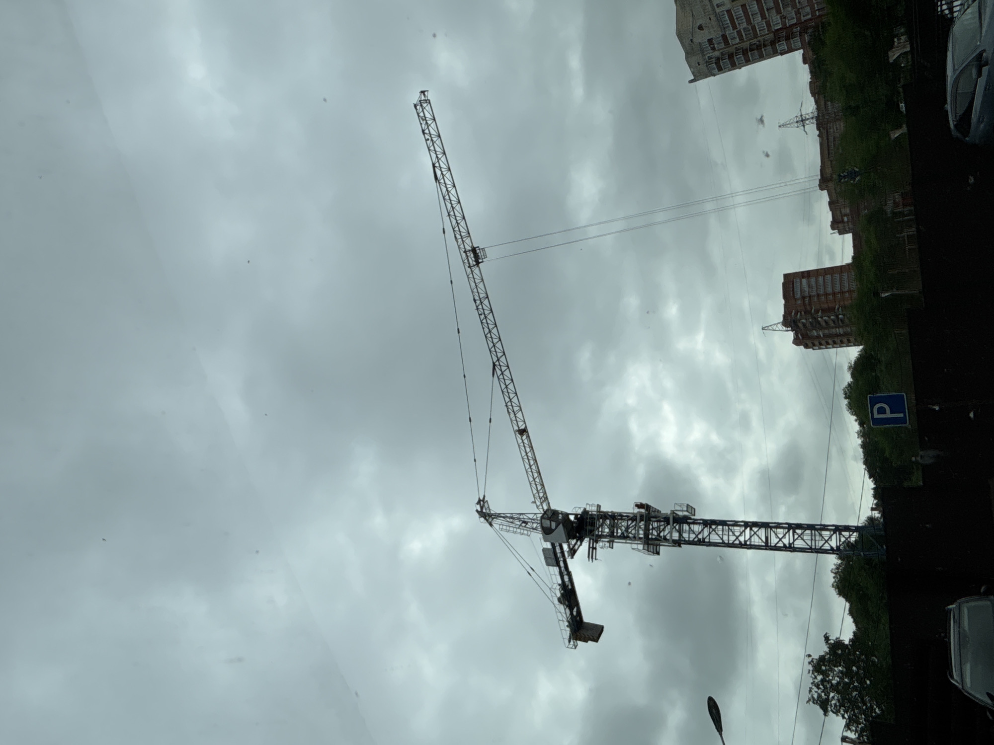 The image captures a construction crane against a cloudy sky. In the background, several residential buildings and trees are visible. A blue parking sign is mounted on a fence in the foreground. The scene appears to be an urban setting, likely in Blagoveshchensk, Russia, judging by the context. No people are clearly identifiable in the image, and no specific actions or interactions are depicted. The overall atmosphere is overcast and somewhat somber due to the cloudy weather.