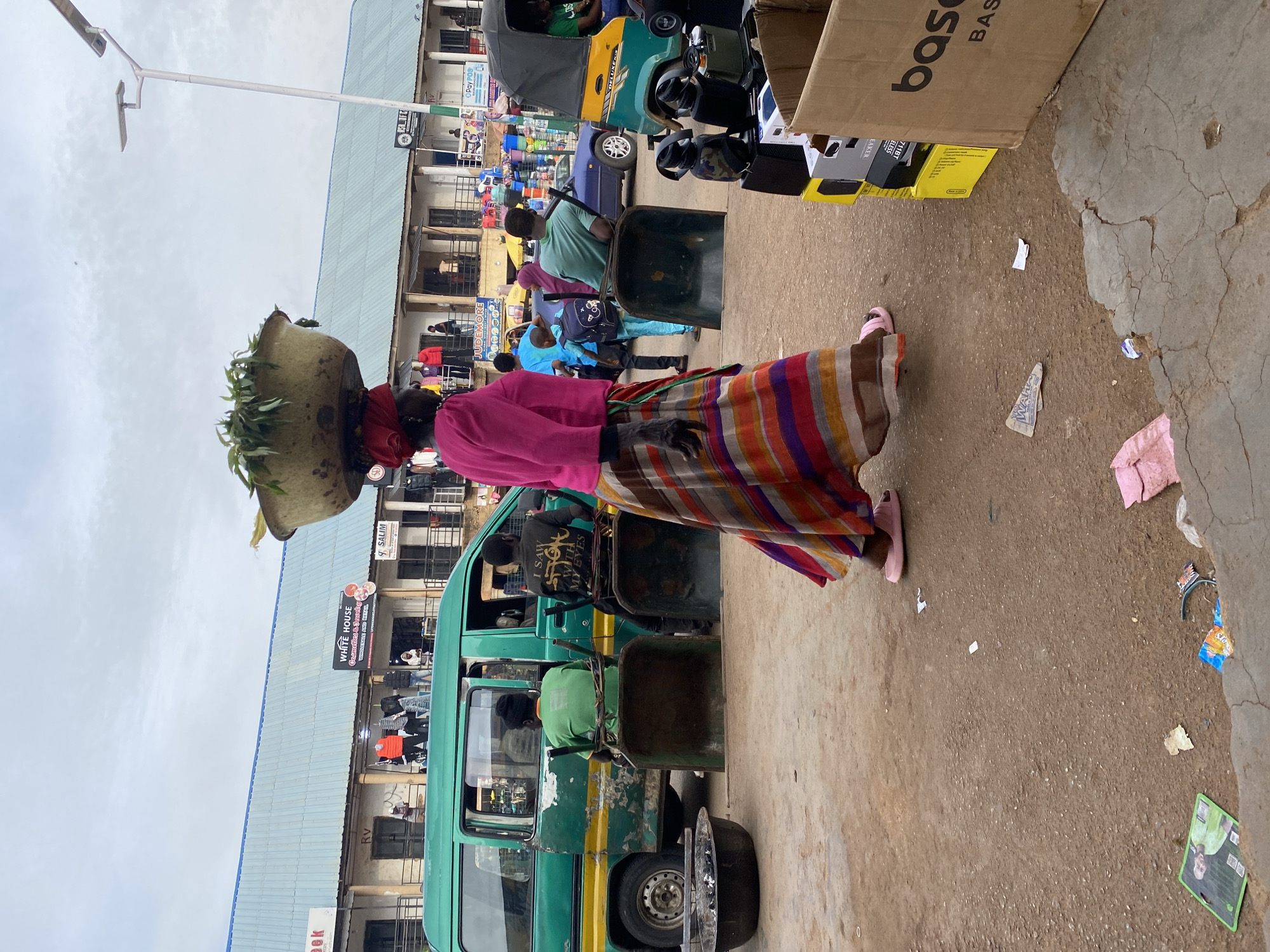 The image shows a bustling street scene in Bukuru, Nigeria. A woman wearing a pink top and a colorful striped skirt is carrying a large basket of greenery on her head. She is walking past a green van with a partially flattened tire.
Behind her, people are milling around in front of a multi-story building with commercial signs and storefronts, including one that reads "WHITE HOUSE Cosmetics & Jewelvag" and "JUDEMORE." There are numerous vehicles on the street, including a yellow and green "DELUXER" tricycle. Various goods, including cardboard boxes and other items, are stacked nearby. The ground is paved, but with cracks and scattered debris. A light pole stands in the background, and the sky is overcast.