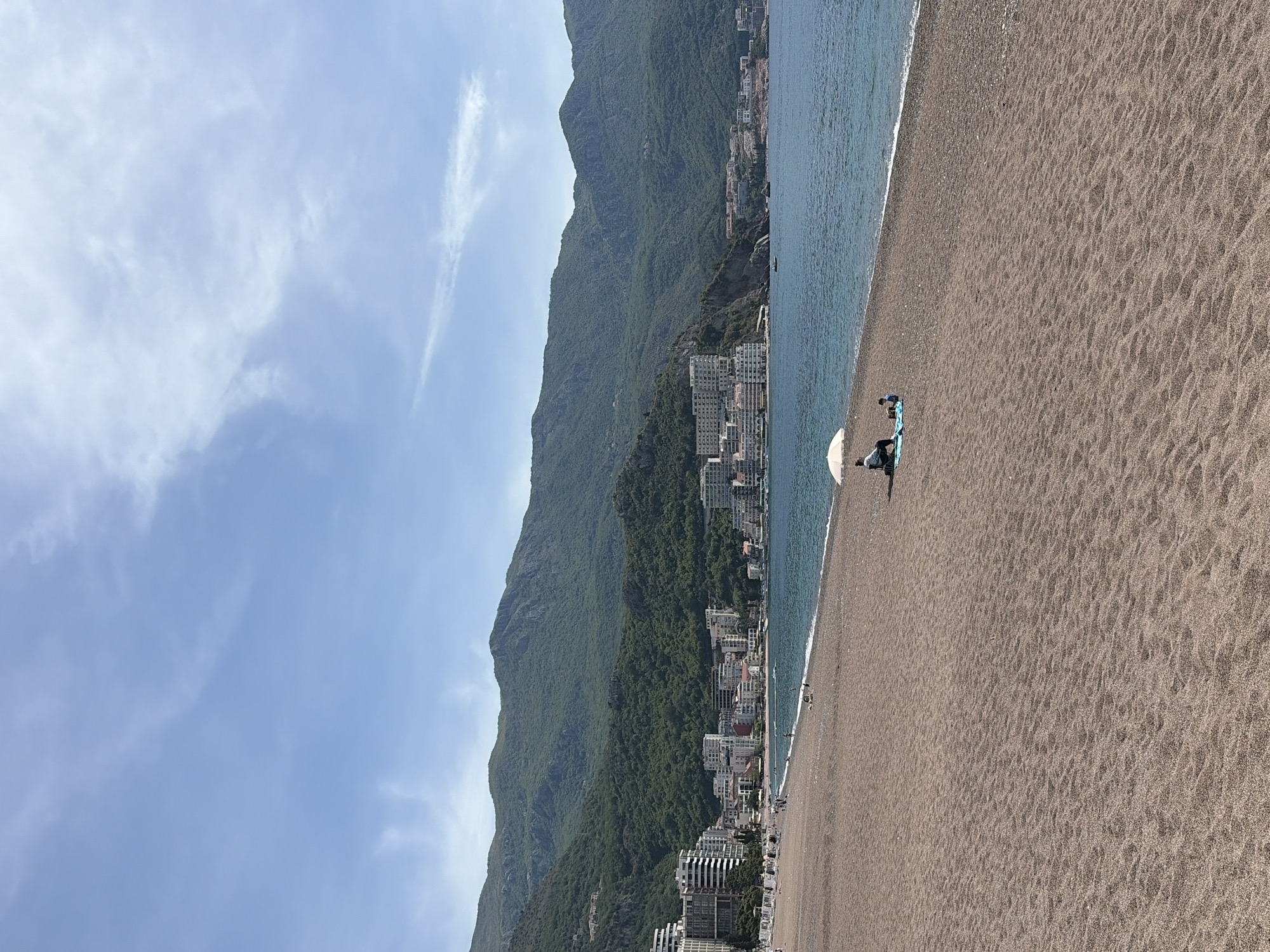 A landscape-oriented photograph depicts a beach along the coast, presumably in Boreti, Montenegro, given the presence of buildings nestled along the mountainside, suggesting a coastal town. The sky is a pale blue with scattered wisps of clouds. A significant portion of the frame is occupied by a mountain range densely covered with green vegetation, positioned to the left. The mountainside is dotted with multi-story buildings, indicative of urban development. A sandy beach fills the lower right of the frame, meeting the blue water’s edge. On the beach, there is at least one person with a beach umbrella. A person is standing next to the umbrella and appears to be interacting with a blue object.