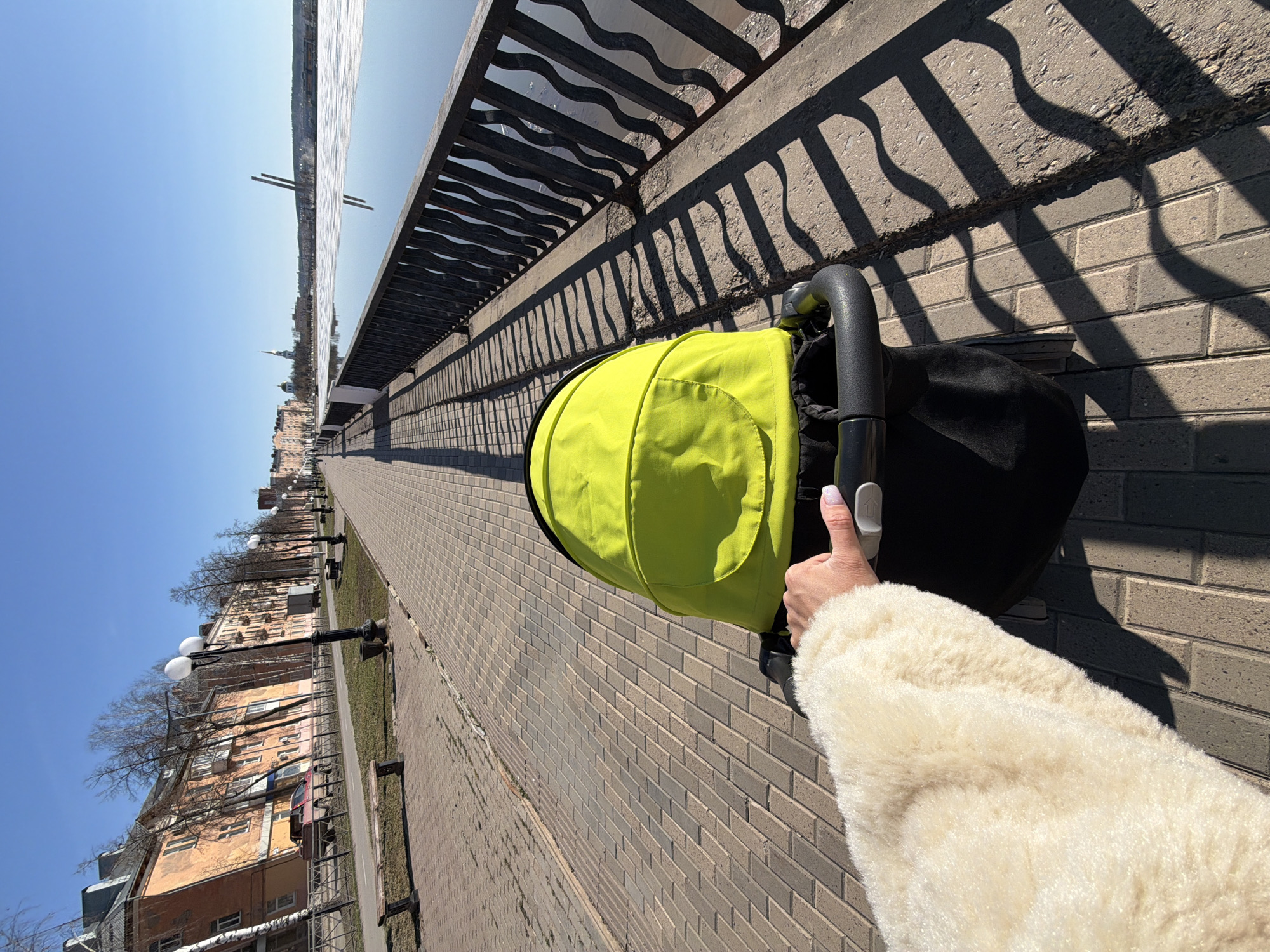 The image captures a first-person perspective of a person pushing a stroller along a sunny embankment in Votkinsk, Russia. The central subject is a bright lime-green and black baby stroller, with a person's right hand and forearm visible, clad in a fluffy, light-colored coat, pushing its handle. The hand has light nail polish.
The scene unfolds on a wide, paved walkway made of light grey rectangular bricks, bathed in strong sunlight that casts prominent, wavy shadows from an ornate dark metal railing. This railing, with decorative undulating patterns, runs alongside the walkway, separating it from a vast, calm body of water—likely the Votkinsk Pond. The water's surface reflects the clear, bright blue sky, stretching towards a distant skyline where old multi-story buildings and a slender spire are visible. Two dark, vertical structures stand prominently in the water.
On the other side of the walkway, a line of residential buildings with an older architectural style can be seen, partially obscured by bare trees. Classic streetlights with spherical white lamps are interspersed along this stretch. The clear blue sky and sharp shadows indicate a bright, sunny day, likely during mid-morning or afternoon. The bare trees suggest the season is late winter or early spring. The overall activity is a leisurely outdoor stroll along the waterfront. No visible text is present in the image.