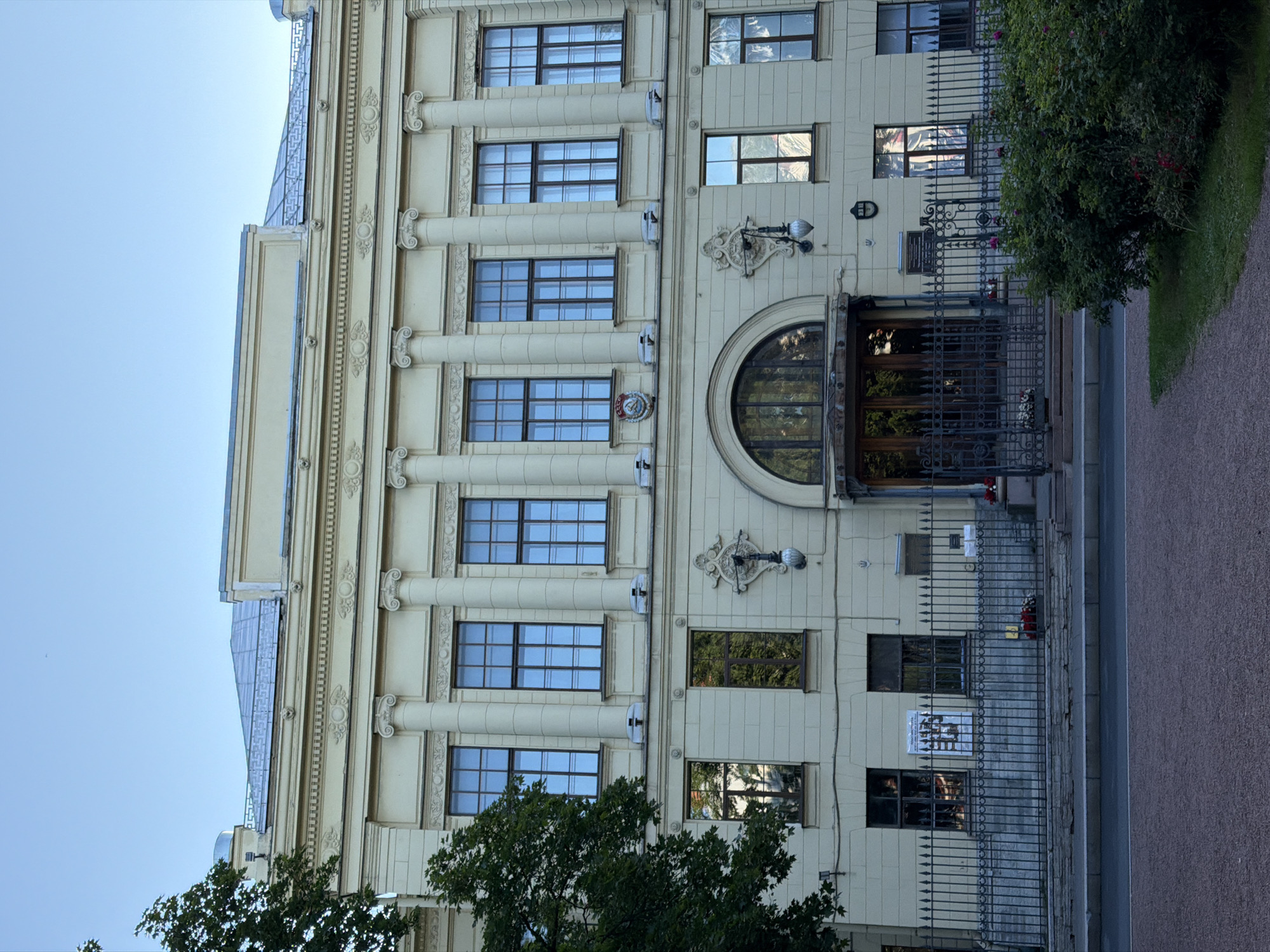 The image depicts the facade of a grand, light yellow building in Saint Petersburg, Russia, likely a public institution or historical landmark. The architecture features classical elements such as columns, ornate moldings, and a symmetrical arrangement of large, multi-paned windows. The building has a light-colored, possibly metal, mansard roof.
In the foreground, a dark metal fence with pointed finials encloses a pathway or garden area. Beyond the fence, a dark green bush with red and pink flowers is prominent on the right side of the image. A paved walkway, possibly a road or sidewalk, runs across the bottom of the frame.
The building's entrance is a large, arched doorway with heavy wooden doors and a decorative metal grille. Above the entrance, an ornate lamppost is mounted on the wall. Windows on the lower floor reflect the surrounding greenery and sky. A sign with Cyrillic text is visible near the entrance. A crest, appearing to be Soviet-era, is mounted between windows on the upper floor.
A large tree with dense green foliage is visible on the left side of the building, partially obscuring the facade. The sky above is clear and blue. The overall scene suggests a serene daytime setting in a well-maintained urban environment. There are no people visibly present in the image.