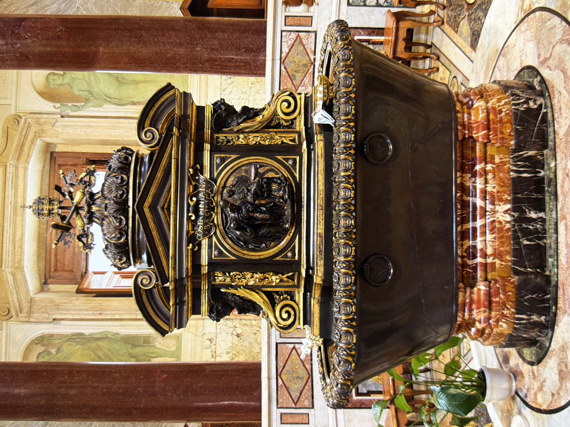 The image shows a large, ornate baptismal font in a church. The font is made of dark bronze with gold accents and is decorated with intricate carvings. The base of the font is made of marble with red and white veins. A small potted plant with green leaves sits to the left of the font. In the background, there are large red columns and frescoes on the walls. The floor is made of patterned marble tiles. There are no people visible in the image. This scene is not located in Kyiv, Ukraine, but rather in St. Peter's Basilica in Vatican City.