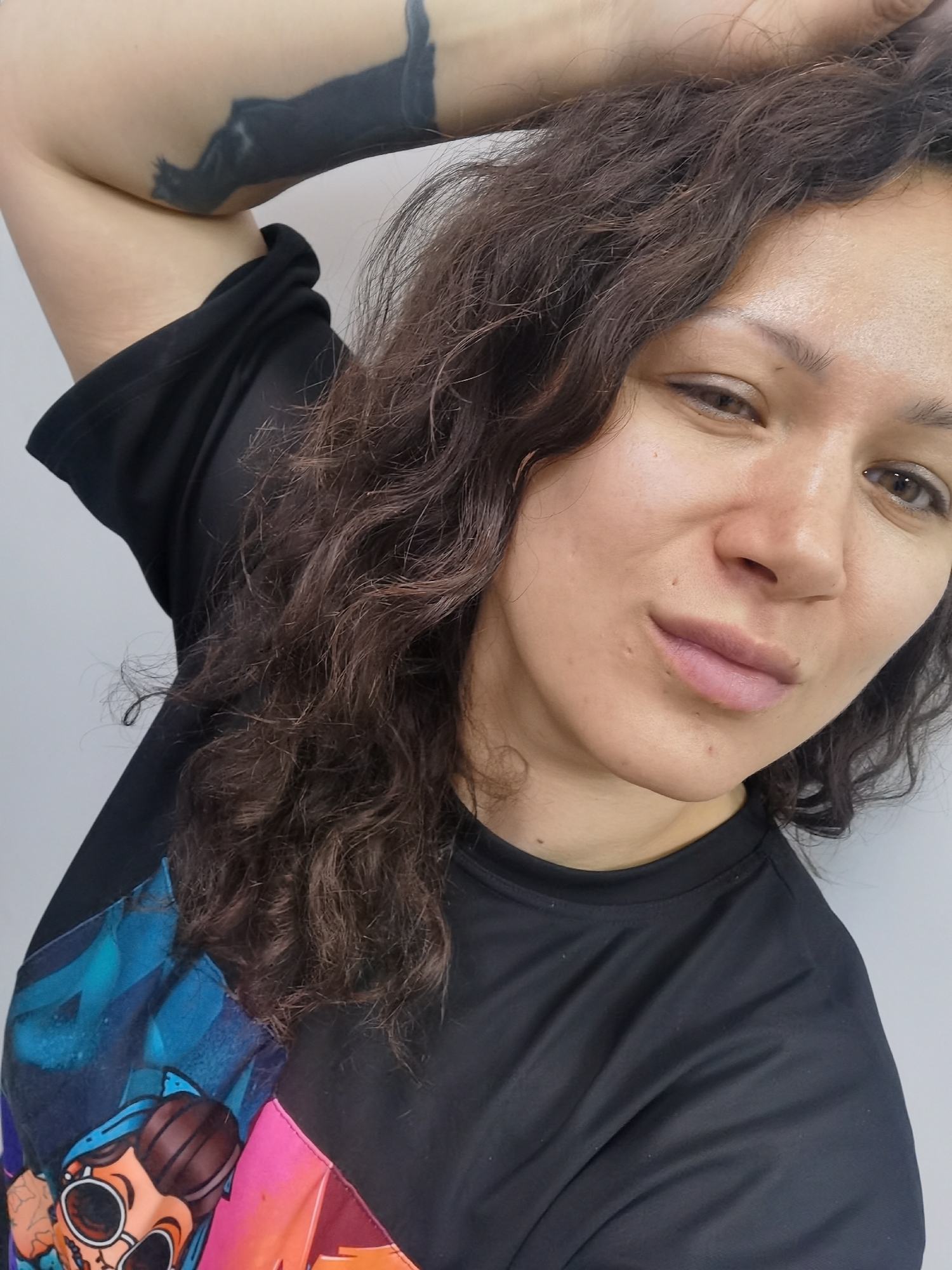 A selfie of a woman with curly brown hair wearing a black t-shirt with a cartoon graphic on the shoulder. She has a tattoo on her raised left arm. The background is a plain white wall. The location is not discernible as Chaikovsky, Russia, based on the provided image.