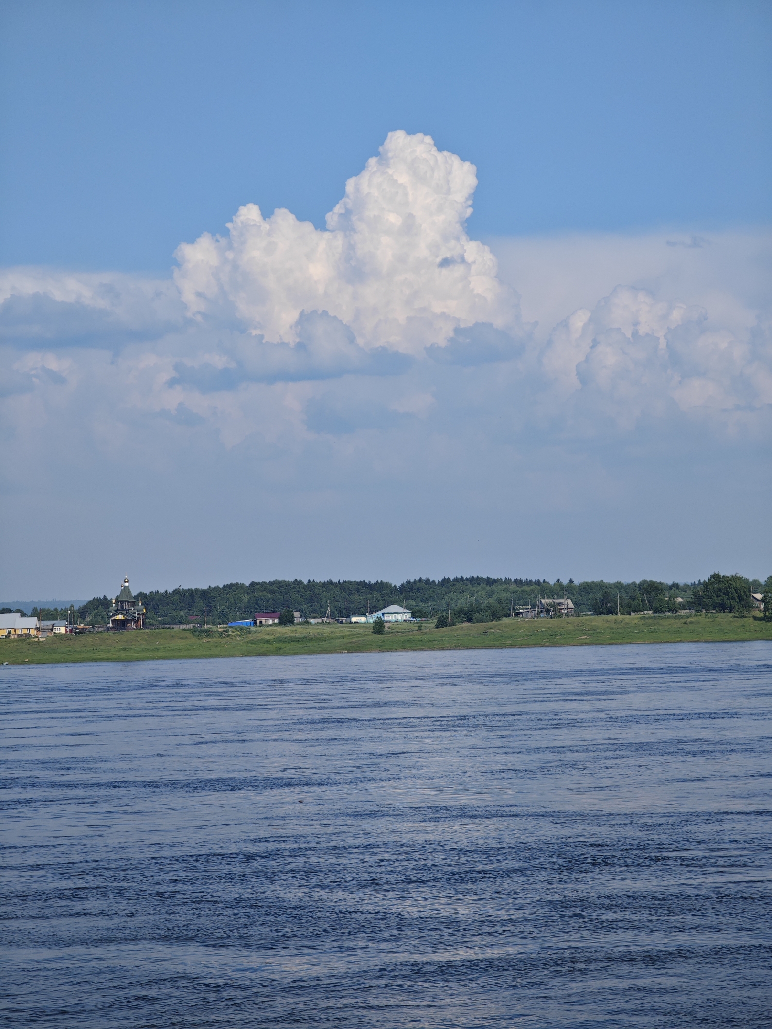 A wide, blue river flows in the foreground, its surface rippled by the wind. Beyond the river, a lush green grassy bank stretches across the scene, dotted with trees. Further inland, a cluster of buildings forms a small settlement. These include several houses, some with red roofs, and a prominent, dark wooden church with a golden dome, situated on a slight elevation. A dense forest forms the backdrop to the settlement. The sky above is a clear blue, marked by a large, puffy white cloud formation. The image appears to capture a serene rural landscape in Verkhnepashino, Russia.