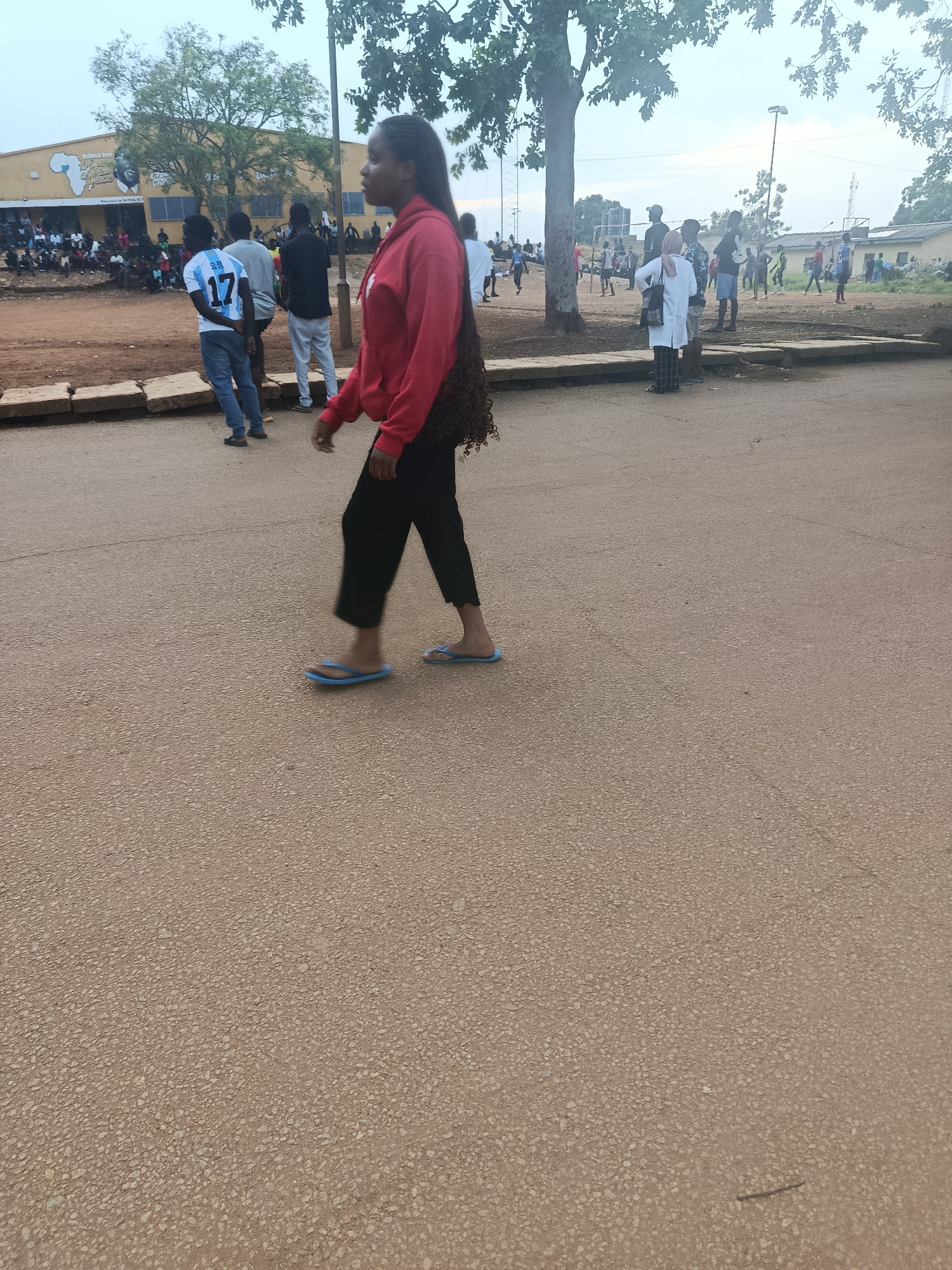 The image depicts an outdoor scene, likely in Rafin Par, Nigeria, with a mix of people and structures. In the foreground, a young woman with dark braided hair is walking towards the left of the frame. She is wearing a red hooded sweatshirt and black pants, and blue flip-flops. Her right hand is extended slightly forward, and she appears to be looking to her left.
In the middle ground, there is a group of people gathered, some seated and some standing. A building with a yellow facade and blue trim is visible in the background, featuring a mural with text and an image. Trees with green foliage are scattered throughout the scene, with one large tree dominating the left side of the frame.
Further in the background, more people are visible, some appearing to be engaged in activities or conversation. The ground is predominantly paved with a coarse, light brown material. The overall lighting suggests it is daytime, possibly late afternoon given the softer light.