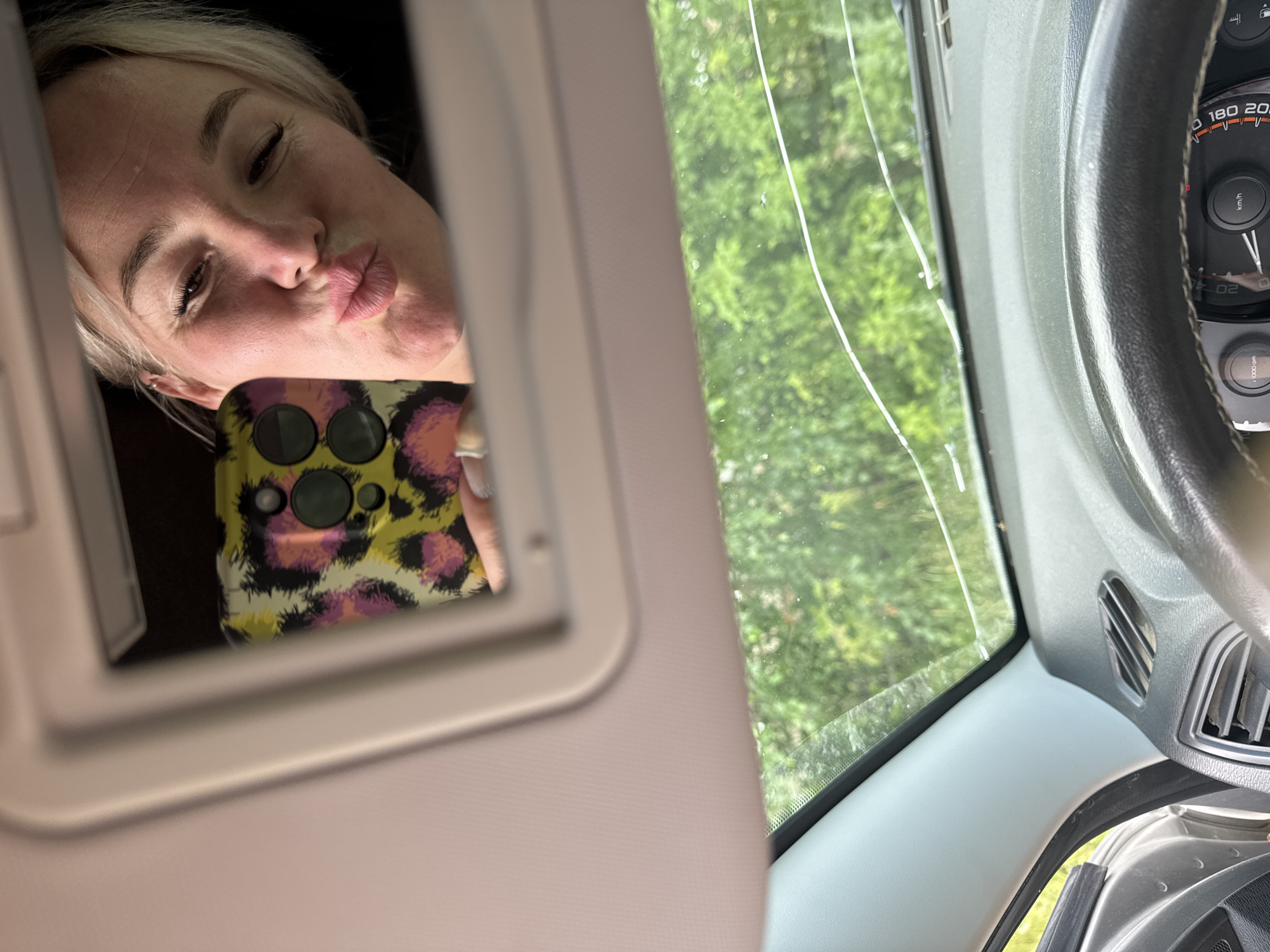 A woman is taking a selfie in the mirror of a car's sun visor. She has blonde hair and is puckering her lips for the photo. Her phone, with a leopard print case, is visible in the reflection. The car's interior, including the dashboard and steering wheel, is partially visible. Outside the car, green trees are seen through the windshield. The image was taken in Nozhovka, Russia.