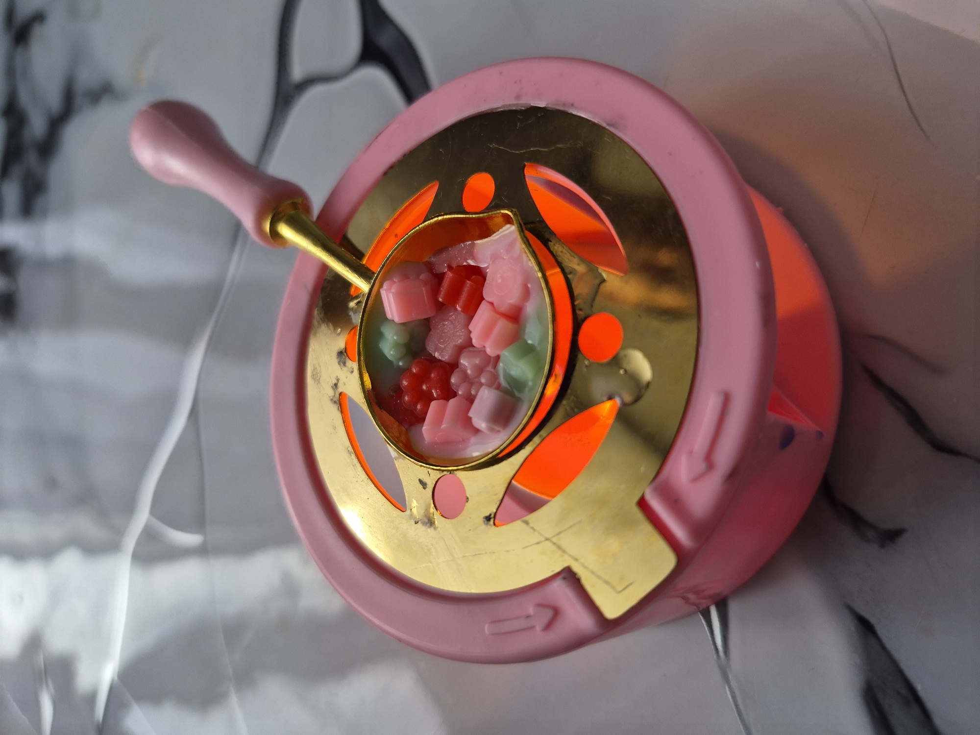 The image shows a pink and gold wax seal warmer. Inside the gold melting spoon, which is part of the warmer, there are small, colorful wax beads in shades of pink, red, and green. The warmer is placed on a surface with a marble-like pattern. The image does not provide any information related to the city of Tobolsk, Russia, or any other location. The scene primarily focuses on the wax seal warmer and its components. There are no people present in the image.
