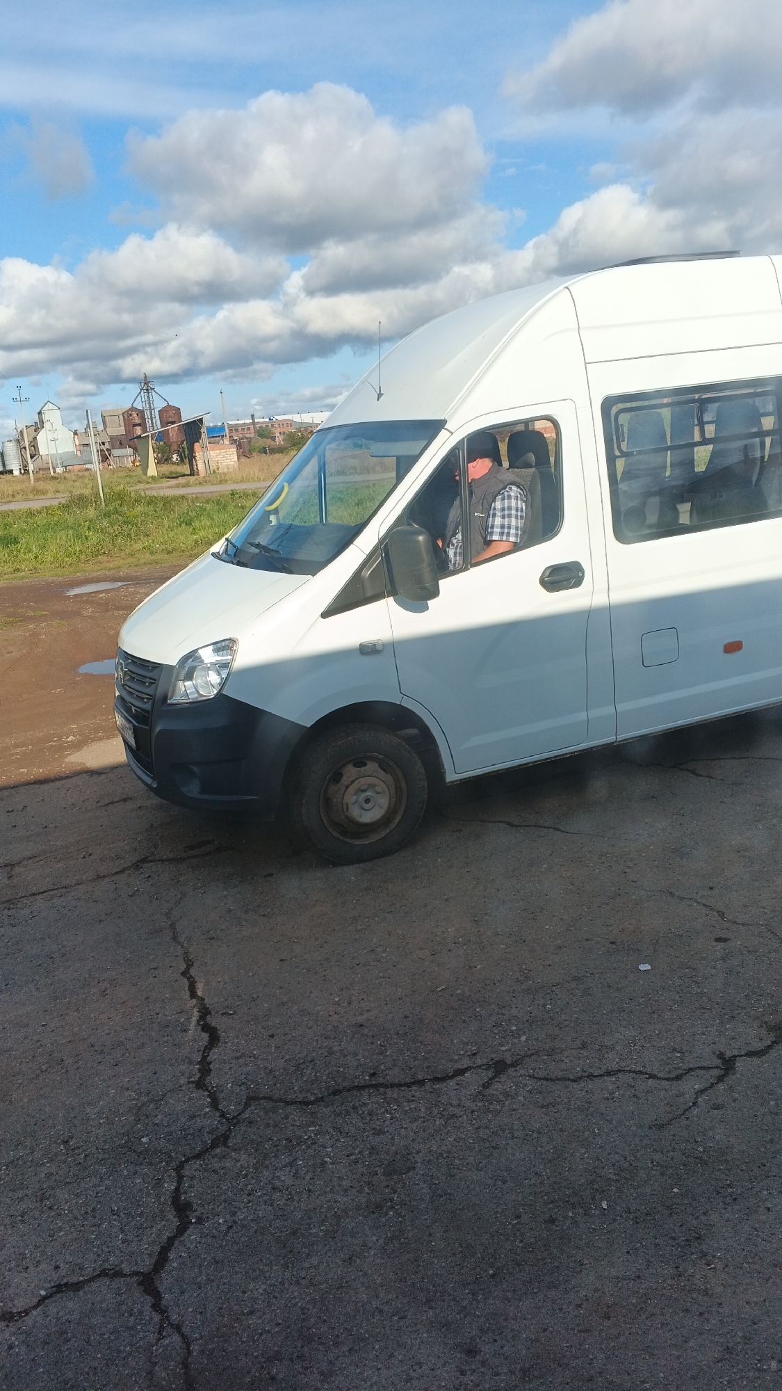 A white Gazelle minibus is parked on cracked asphalt. A person wearing a plaid shirt and a dark vest is visible in the driver's seat. In the background, there are industrial buildings, a fence, and grassy areas under a partly cloudy blue sky. The scene appears to be in an industrial or rural area.