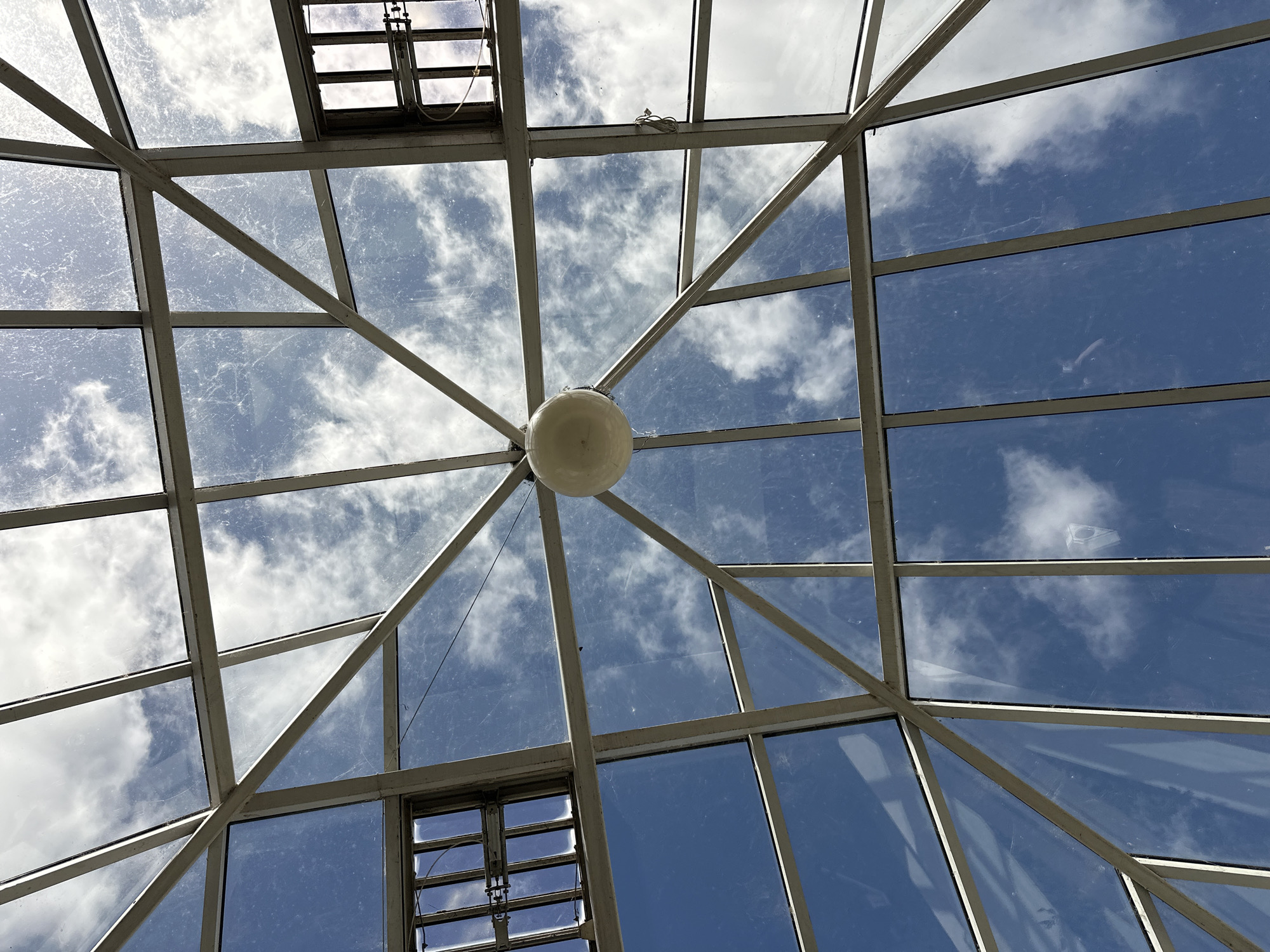 The image is a low-angle shot looking up at a glass-paneled ceiling with a metal framework. The sky, visible through the glass, is blue with scattered white clouds. There are two square vents with metal grilles in the upper corners of the frame. A round, off-white light fixture hangs from the center of the ceiling structure. The glass panes appear to have some dust or streaks on them. The scene is an interior view of a building's ceiling, looking outwards to the sky. The location in Carlow, Ireland, is not discernible from the image itself.