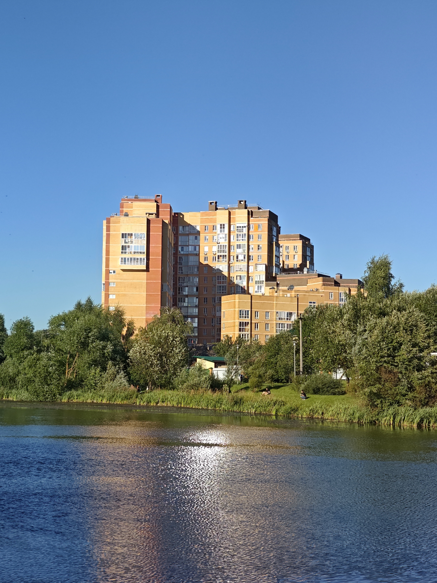 The image captures a scene in the city of Shcherbinka, Russia, featuring a large, modern residential building complex situated behind a tranquil body of water. The complex is characterized by its multi-story brick structures, primarily in shades of orange and yellow, with numerous windows and balconies. Lush green trees and reeds line the bank of the water, creating a natural buffer between the buildings and the water's edge. The water itself reflects the blue of the clear sky and the warm hues of the buildings, with shimmering sunlight creating a textured surface. In the foreground, the calm water dominates the lower half of the frame. The middle ground shows the verdant bank with the trees and the residential buildings beyond. In the background, the expansive, cloudless blue sky fills the upper portion of the image. A few small figures are visible on the grassy bank, appearing to be relaxing or enjoying the outdoors. The overall impression is one of a peaceful urban environment integrated with natural elements.