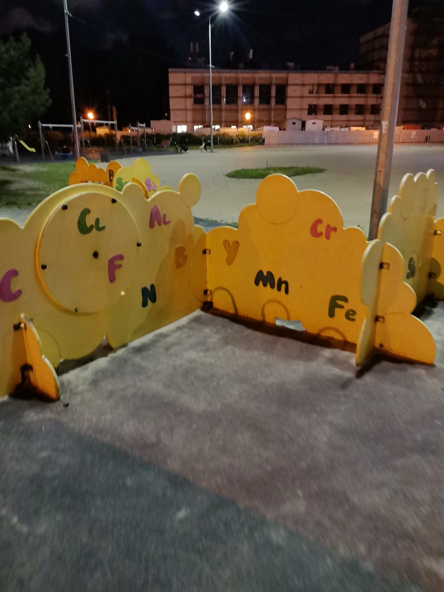 This image shows a children's playground at night in Kazan, Russia. In the foreground, a yellow playground structure with cloud-shaped elements is visible. The structure has various letters and symbols on it, including "C", "Cl", "F", "N", "Al", "B", "Y", "Mn", "Cr", and "Fe". The ground is a mixture of dark pavement and a lighter, sandy surface. In the background, there are swing sets, a slide, and a multi-story building under construction or renovation. The scene is lit by streetlights and artificial lighting, creating a dark and slightly eerie atmosphere. There are two dogs in the mid-ground, one black and white and the other dark-colored, appearing to be interacting in the open space of the playground.