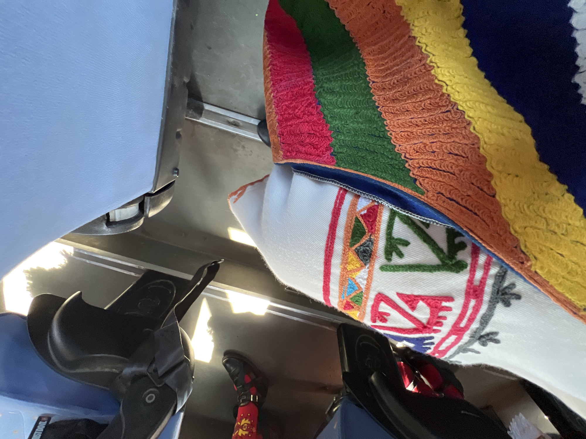 The image displays an interior scene, likely within a public transport vehicle such as a bus or train, viewed from a slightly elevated perspective looking downward. The specific location in the city of Slivovo, Bulgaria, cannot be confirmed or determined from the visual evidence provided.
To the upper left, a portion of a blue seat is visible. Below it, a black, multi-component mechanism, possibly a seat adjustment lever or part of a footrest, is present. Further down and to the left, a person's left foot, clad in a black shoe or boot with red accents, is visible on a dark floor. The leg above the shoe is covered by a vibrant, red fabric patterned with yellow designs.
In the foreground, a person's lap or lower body is covered by two distinct fabric items. The top item is a white textile, possibly a blanket or garment, featuring intricate embroidery in horizontal bands. The embroidery includes geometric motifs and stylized letters or symbols in red, orange, yellow, blue, and green. Below or adjacent to the white textile, a colorful, horizontally striped fabric is visible, likely a blanket. The stripes, which appear to be knitted or crocheted, are distinctly colored in dark blue, yellow, orange, green, and red, arranged from bottom to top in the visible section.
The floor beneath is dark and reflective, showing bright rectangular reflections of light sources, possibly windows, from the upper left. A metal rail or strip runs horizontally across the floor, consistent with vehicle interior construction. To the upper right, a light blue, textured surface, likely the back of another seat, is visible.