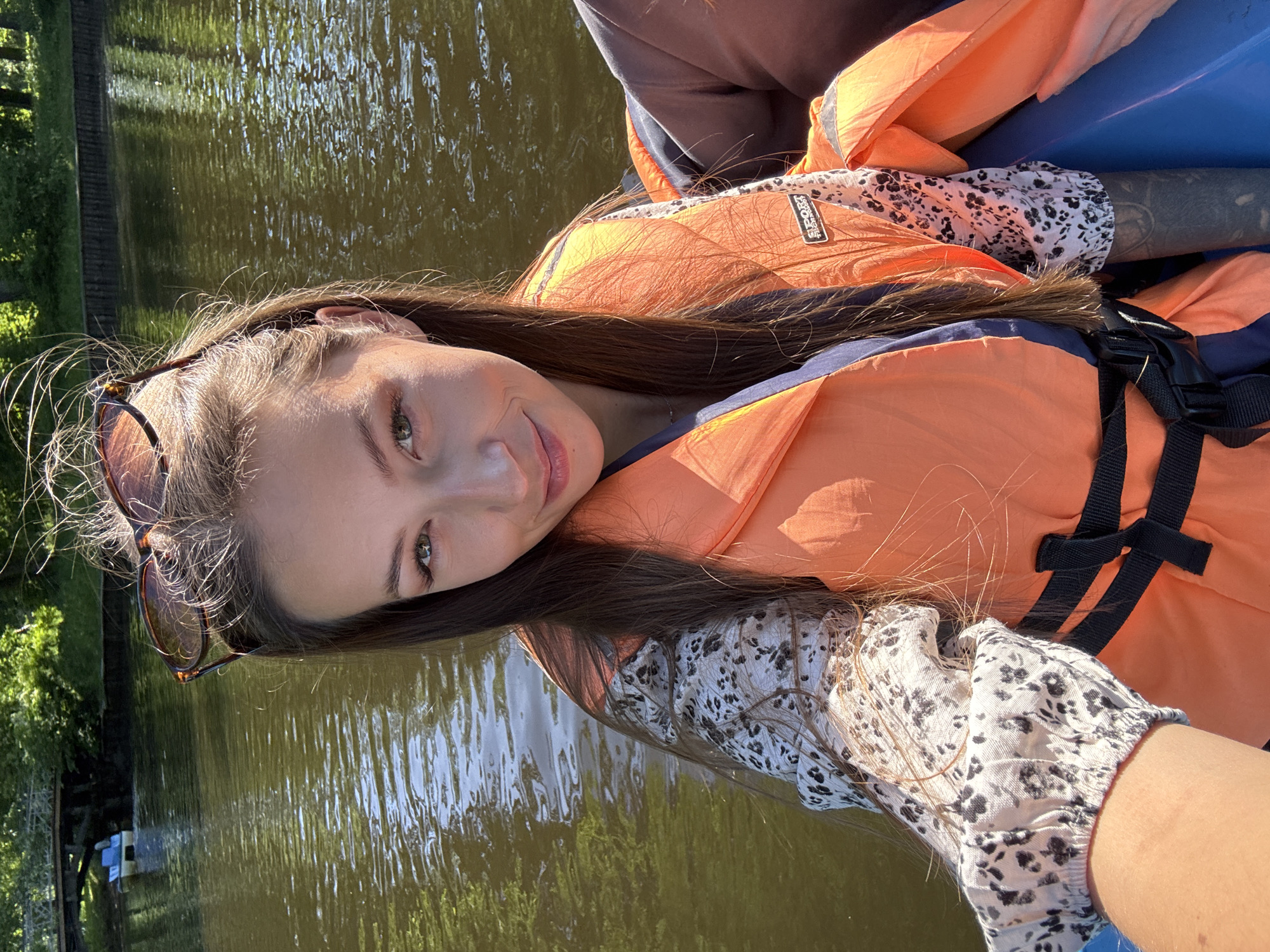 A young woman is pictured outdoors on a sunny day, engaged in a water activity, likely kayaking or boating, in Saint Petersburg, Russia. She is wearing an orange and dark blue life vest over a long-sleeved white shirt with a speckled or small floral black/dark gray pattern. Her long, dark brown hair flows over her shoulders, and sunglasses are pushed up onto her head. She has fair skin, light-colored eyes, and a soft, pleasant smile directed towards the viewer, suggesting a selfie or close-up shot. The background features calm, reflective water, indicating a river or canal, with hints of green foliage along the banks. Bright sunlight creates reflections on the water and highlights her hair and parts of the life vest. A black label with white text reading "SPORT" is visible on her life vest. A blue object, possibly part of the watercraft, is also visible. The overall mood is relaxed and recreational.