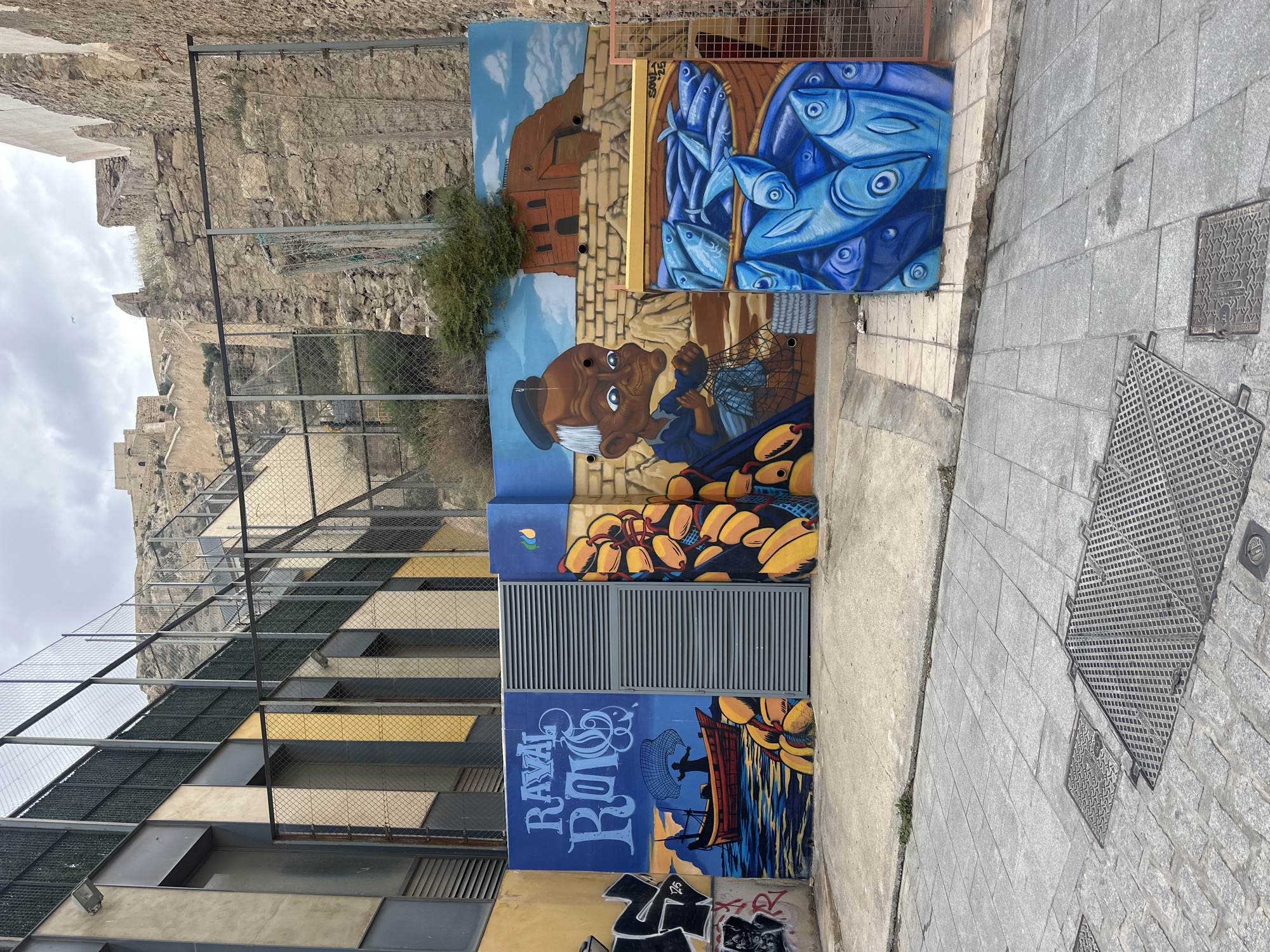 The image shows a colorful mural painted on a building in an urban setting. The mural depicts various maritime-themed scenes, including a fisherman with a net, a basket of blue fish, and a boat on the water. The name "Rayal Roig" is prominently displayed on one of the panels. In the background, a stone castle or fortress is visible on a hill. A fence runs along the side of the building with the mural. The location is Unknown, Unknown.