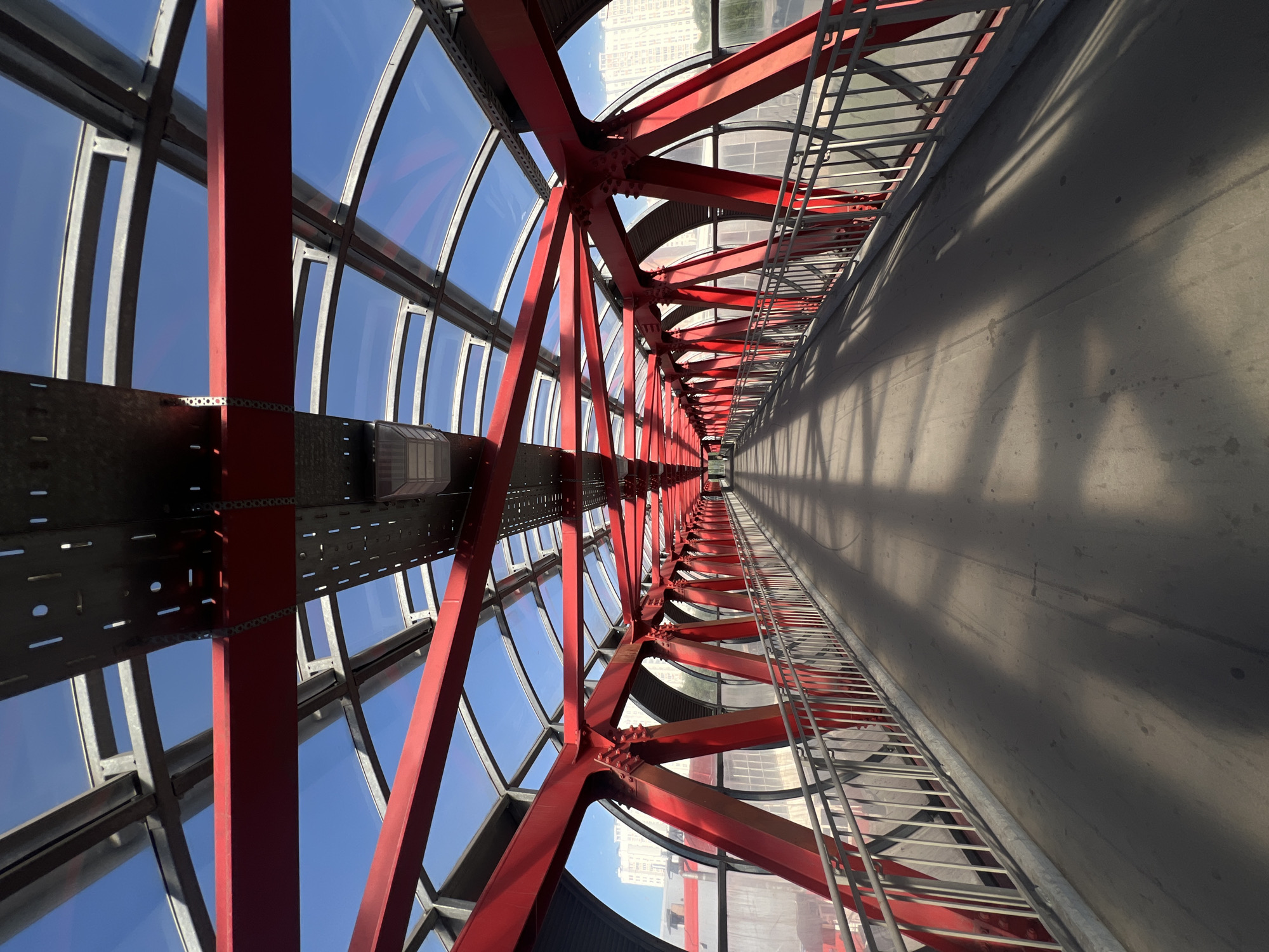 The image shows a pedestrian overpass in Moscow, Russia. The overpass has a red metal frame and a glass roof, allowing sunlight to filter through and cast shadows on the walkway. The walkway is made of concrete and has a metal railing on either side. In the background, there are buildings, suggesting an urban environment. The sky is clear and blue. The perspective is from the middle of the overpass, looking down its length towards a vanishing point. There are no people visible in the image.