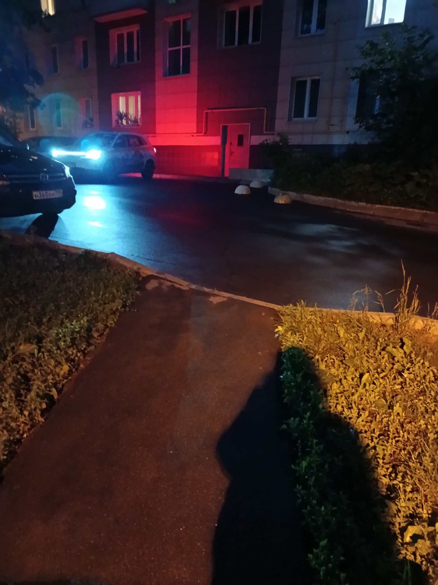 The image shows a street scene at night, likely in a residential area of Moscow, Russia, based on the license plate of the black car visible in the foreground. Two cars are parked on the street, with their headlights illuminating the wet pavement. A multi-story building is visible in the background, its facade reflecting red light, possibly from streetlights or emergency vehicle lights. Shrubs line the edge of the pavement along the walkway. The shadows of a person are visible, suggesting someone is standing nearby or walking through the scene.