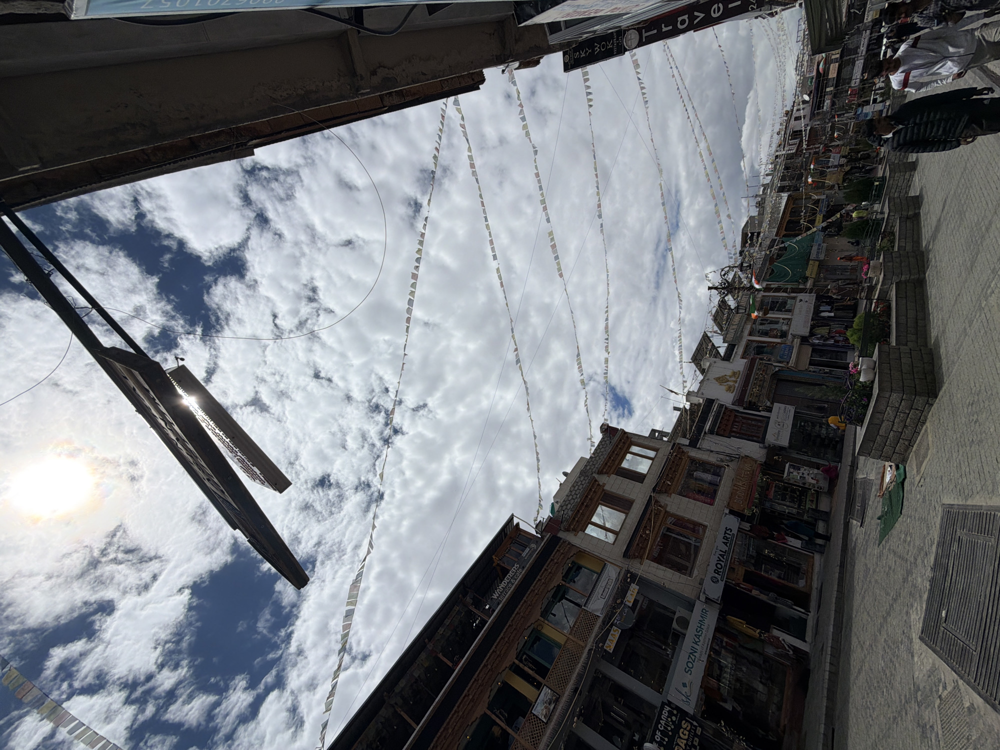 This is a low-angle, wide shot of a street in Leh, India. The sky is filled with white, puffy clouds and the sun is bright, with rays filtering through. Prayer flags are strung across the street, some catching the sunlight. The buildings lining the street are multi-story structures with traditional Tibetan architecture, featuring wooden balconies and ornate windows. Shops and cafes are visible at street level, with some signs indicating businesses like "Wanderers Terrace Cafe" and "Royal Arts". People are walking on the street, some in traditional attire. The street is paved with stone and there are manhole covers visible. A dark, angled structure hangs from above, possibly a sign or part of a building. The overall scene suggests a bustling marketplace or a main thoroughfare in the city.