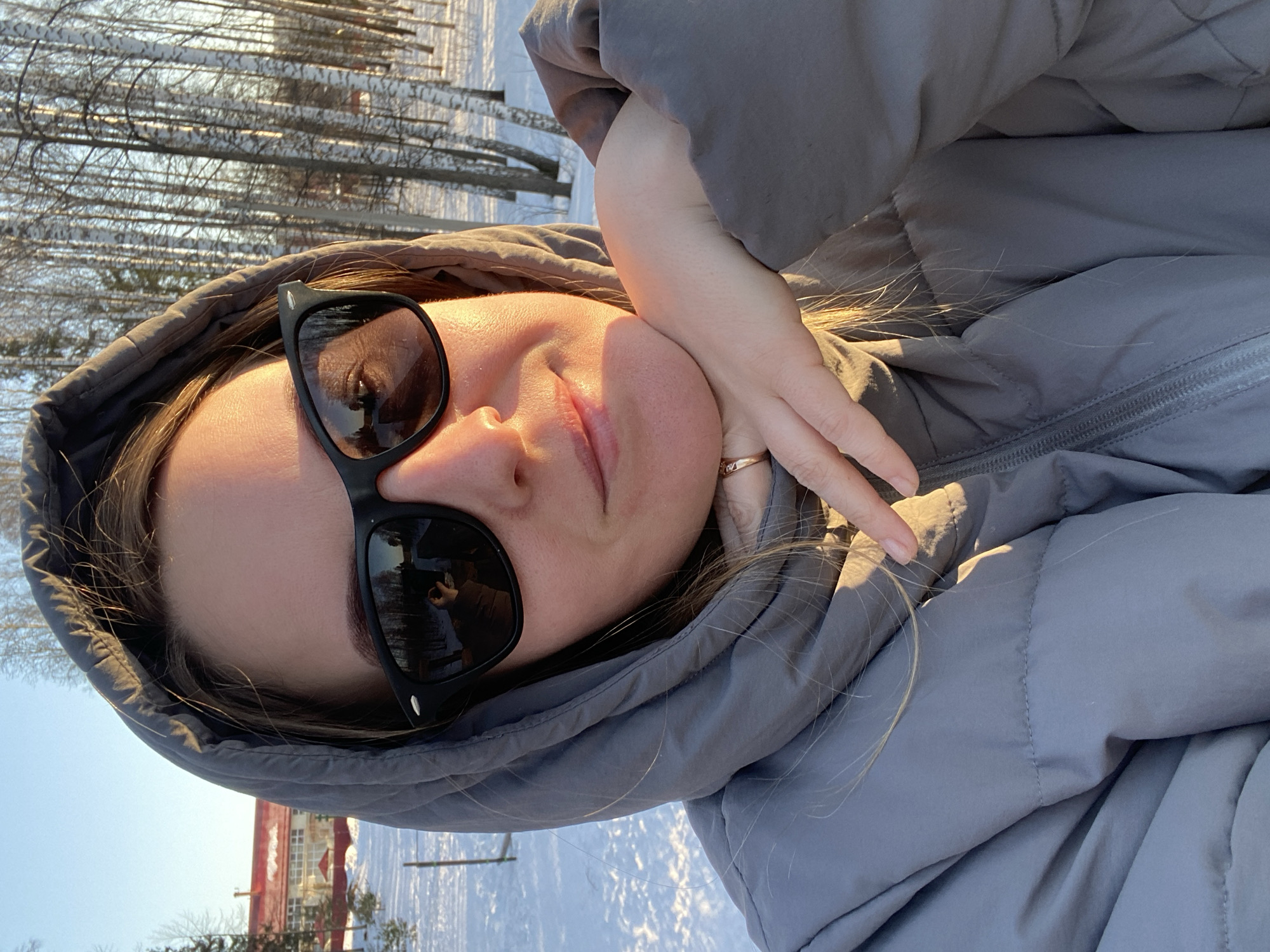 A close-up selfie depicts a female subject in Noviy, Russia, positioned outdoors in a snow-covered environment during daylight. The subject, seen from the mid-chest up, wears a grey hooded puffer jacket with the hood drawn around her face. She is wearing black rectangular sunglasses that reflect a blurred image of an individual (likely the subject herself taking the photo) and surrounding bare trees. Her right hand, adorned with a gold-colored ring on the ring finger, rests against her jawline. Sunlight illuminates the right side of her face and jacket, casting a warm glow. The background features a snow-covered ground. To the left, a light-colored fence post is visible, with a multi-story building featuring a red roof and white facade in the far distance, partially obscured by trees. To the right, numerous bare birch trees with white bark stand in a dense cluster. The sky is clear blue.