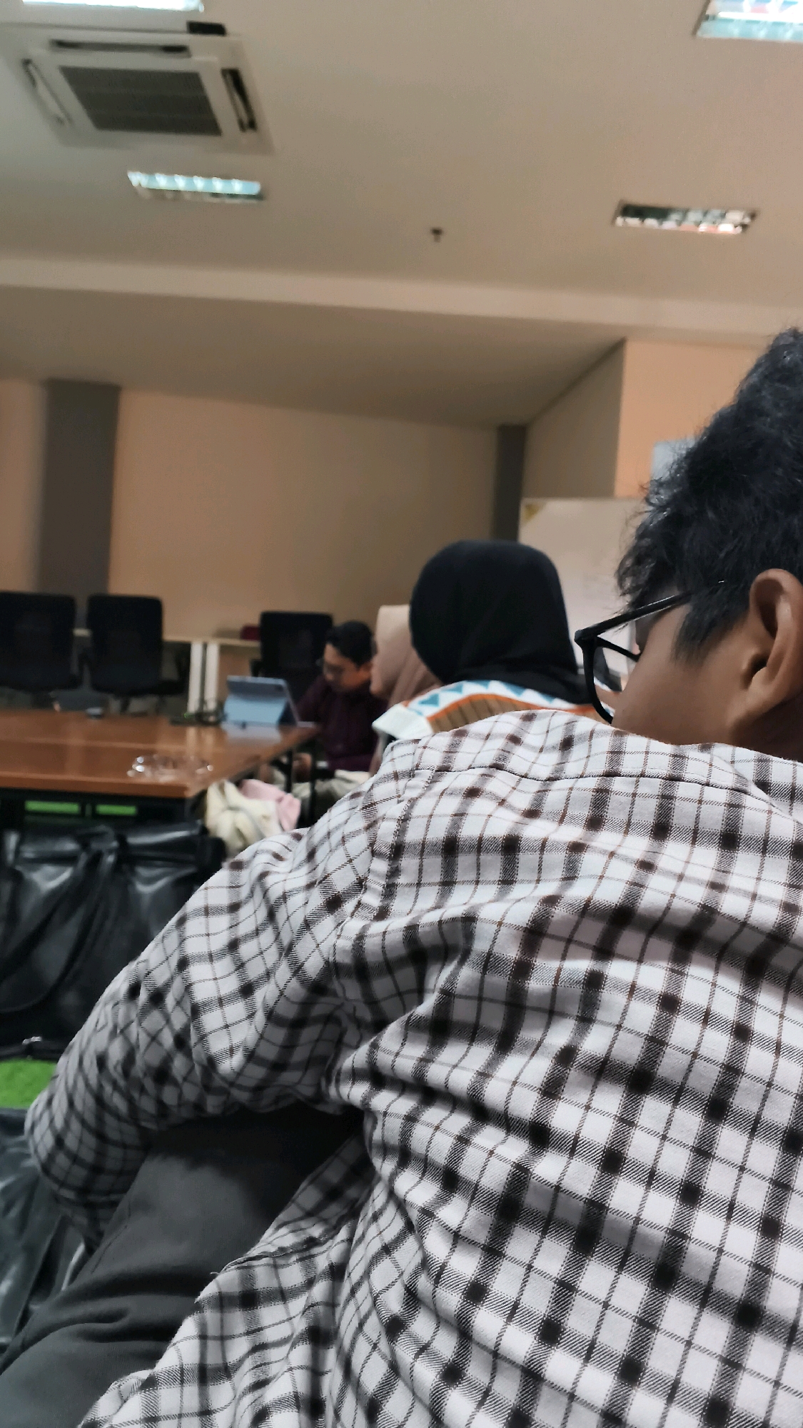 The image depicts an indoor scene, possibly a meeting room or classroom, with a focus on the back of a person wearing a checkered shirt. The person is seated, facing away from the camera, with their right ear and part of their neck visible. They are wearing glasses. In the background, several other individuals are visible, also seated. One person in the mid-ground is wearing a black hijab and a patterned top. Another person further back is wearing glasses and a maroon shirt. The room features a long wooden table, several black chairs, and a projector screen or whiteboard mounted on the wall. The ceiling has air conditioning units and fluorescent lighting. The overall impression is that of a formal gathering or discussion. The image does not provide any visual cues to determine the specific location within Lohbener, Indonesia.