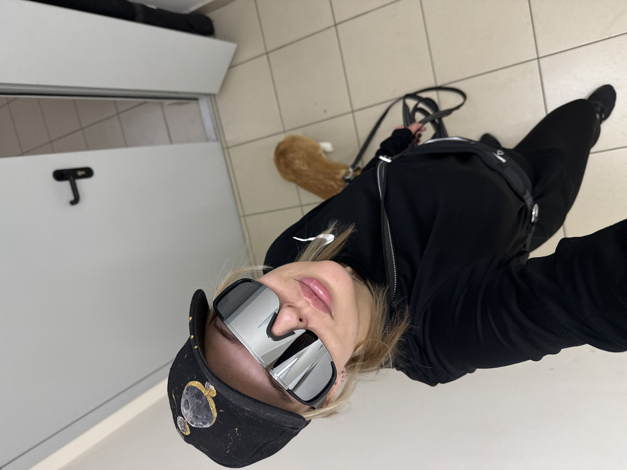 A selfie angled upwards features a person wearing a black baseball cap with celestial objects depicted on it, mirrored sunglasses, and a black zip-up hoodie. The person is wearing black pants and sneakers. A brown and white animal, possibly a dog or cat, is visible behind them. The background appears to be an interior space with light-colored tiled flooring and white walls. A door is also visible on the left side of the frame. The location is possibly in an enclosed room. No specific signs or details confirm a location in Krasnoyarsk, Russia.
