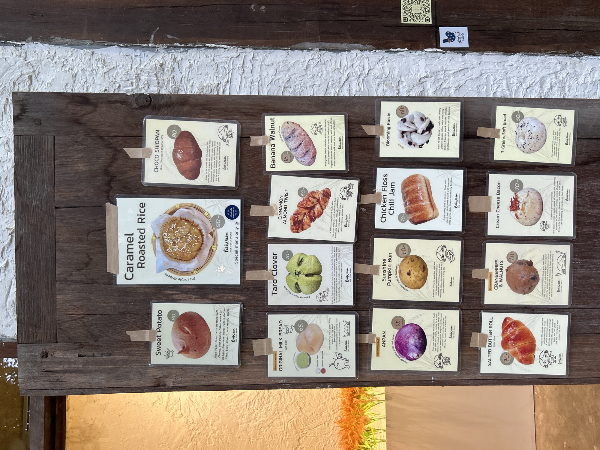 The image shows a menu board for a bakery, likely located in Ban Sai Kham, Thailand, as indicated by a QR code and Thai text on the right side of the image. The menu is displayed on a rustic wooden door, with individual laminated cards advertising various types of bread. Each card features a picture of the bread, its name, a description, and a price. The breads advertised include: Sweet Potato, Caramel Roasted Rice, Choco Shiopan, Original Milk Bread, Taro Clover, Cinnamon Almond Twist, Banana Walnut, Anpan, Sunshine Pumpkin Bun, Chicken Floss Chili Jam, Blooming Raisin, Salted Butter Roll, Cranberries & Walnuts, Cream Cheese Bacon, and 7-Grains Soft Bread. The environment appears to be outdoors, with natural light illuminating the scene and a hint of greenery from plants in the background on the left. The overall presentation is charming and rustic, suggesting a local bakery with handcrafted products.