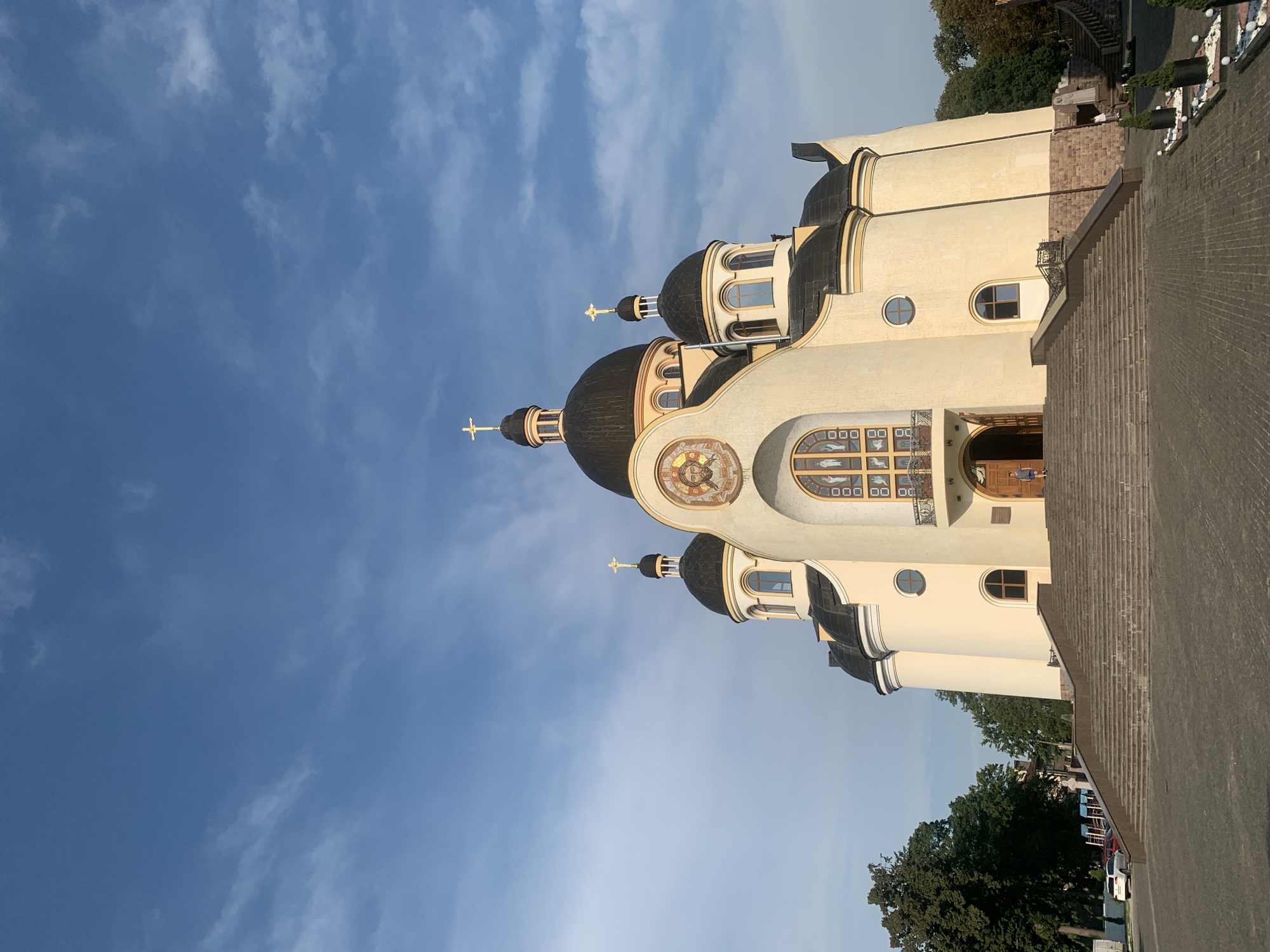 The image depicts the Church of the Transfiguration in Kolomyia, Ukraine, under a bright blue sky with scattered clouds. The church is a prominent, cream-colored building with several dark domes topped with golden crosses. A large, arched stained-glass window is situated above the entrance, flanked by circular windows. A mosaic depicting a face is centered above the large window. A wide set of stone stairs leads up to the main entrance of the church. A few trees are visible on the left and right sides of the building, and there are some decorative plantings in the foreground on the right. A person is visible walking up the stairs on the right side of the entrance, and another person is near the base of the stairs on the right. Several cars are parked in the background on the left. The ground in front of the church is paved with cobblestones.
