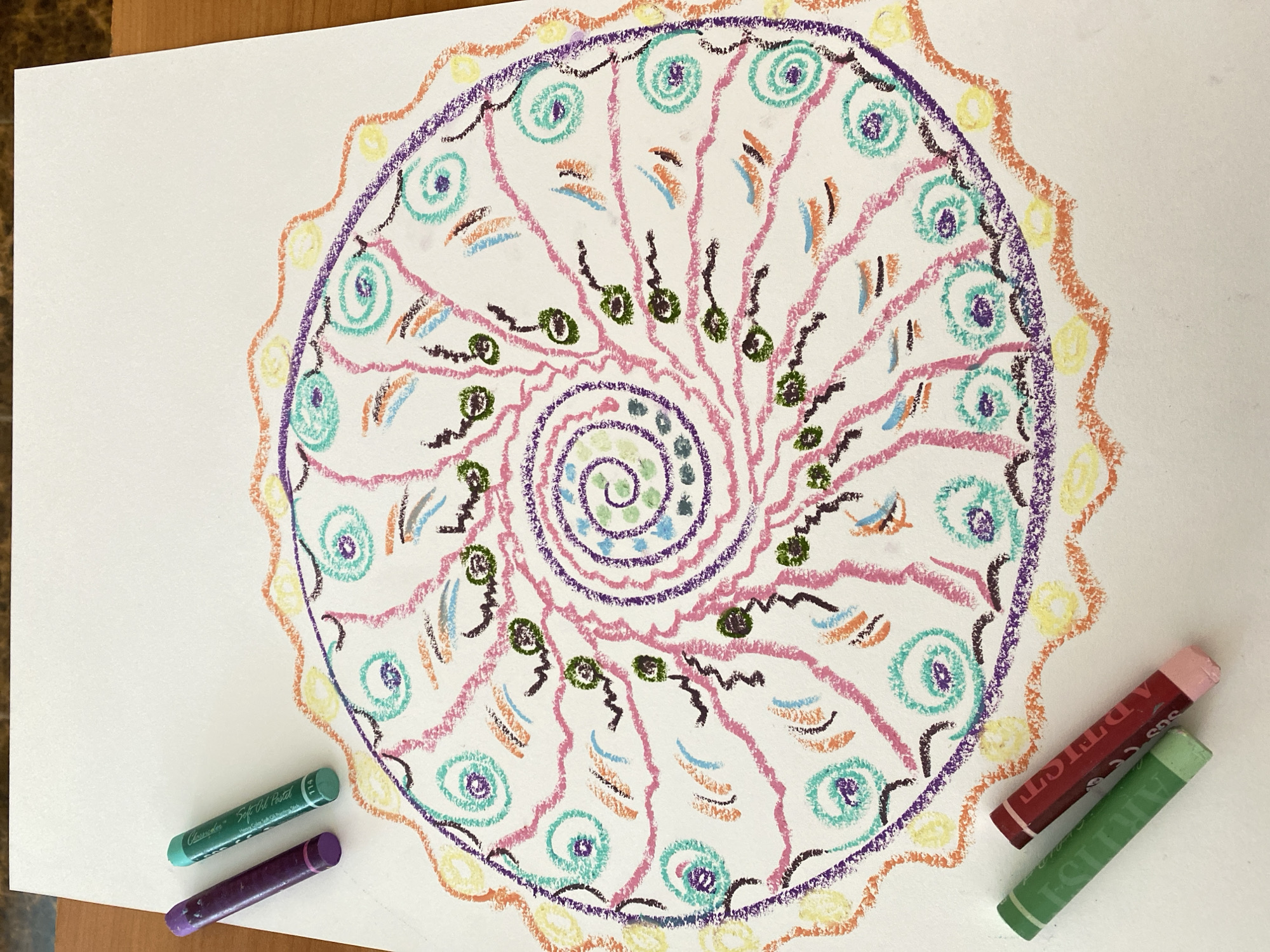 The image depicts a colorful, hand-drawn mandala on a white piece of paper. The mandala is composed of concentric circles, spirals, and various shapes filled with hues of purple, pink, teal, orange, yellow, and green. These elements create a symmetrical, radial pattern. A few pastel crayons in shades of green, purple, and red are scattered beside the drawing on the paper's surface. There is no evidence of the image being in Yekaterinburg, Russia, or any people present.