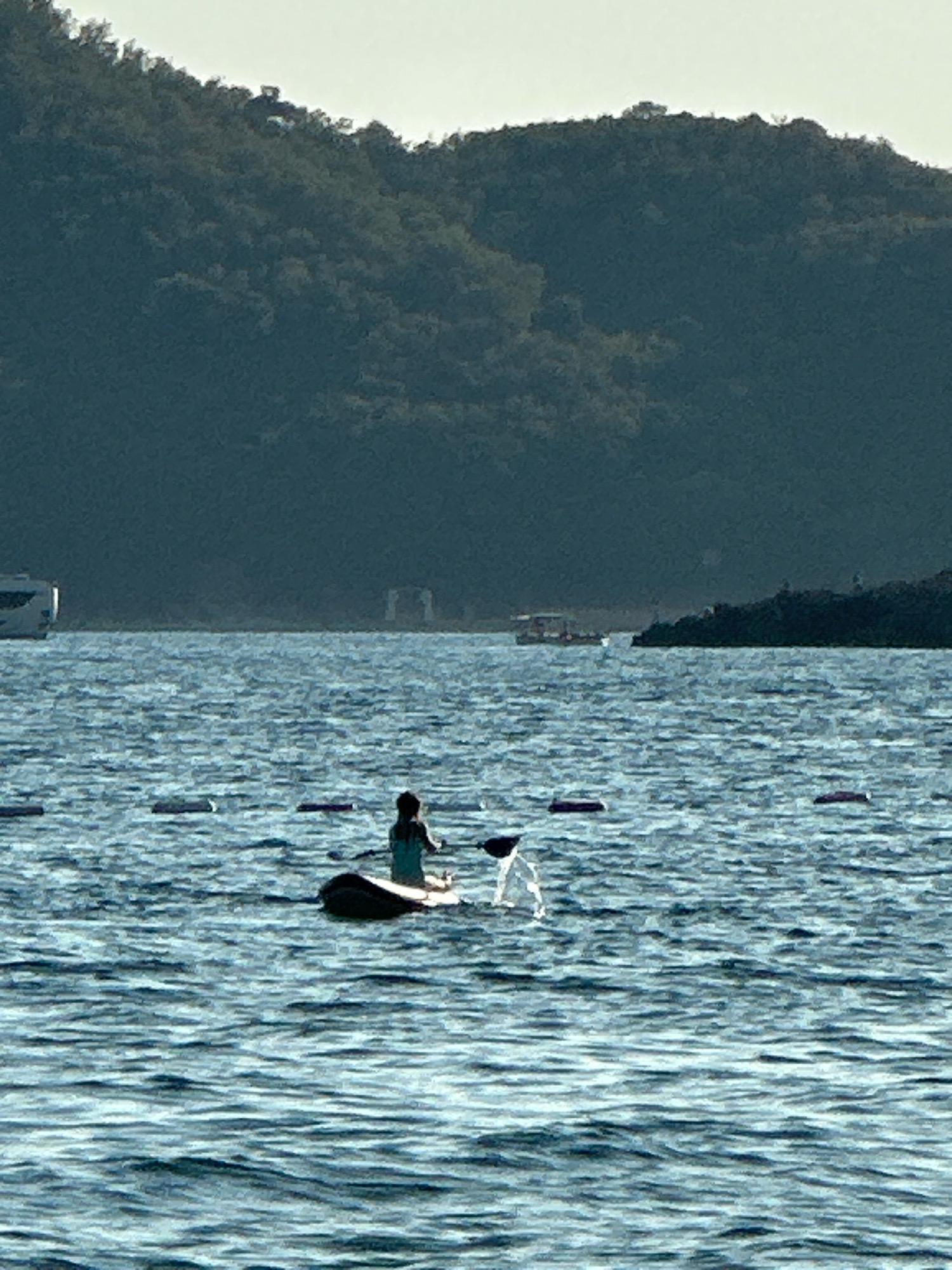 The image depicts a tranquil scene on the water, likely the Bay of Kotor, near Boreti, Montenegro, given the context. In the foreground, a person is on a paddleboard, paddling through calm blue water. The individual is wearing a teal-colored top and appears to be engaged in the activity of paddleboarding. A small splash of water is visible near the paddleboard, suggesting movement. In the background, a dark, tree-covered hillside rises, partially obscuring the sky. A few distant boats and a larger vessel are visible on the water, indicating the presence of other watercraft. The overall impression is one of a peaceful day spent on the water, with the natural beauty of the Montenegrin coastline providing a scenic backdrop.