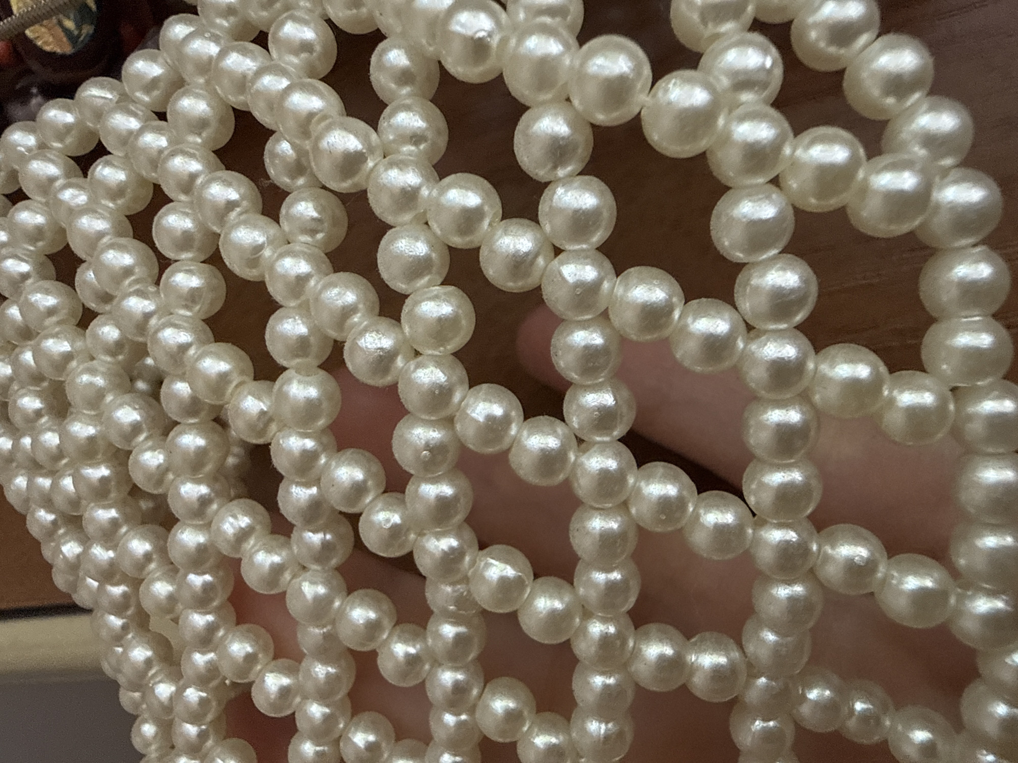 The image displays a close-up view of a pearl-like beaded material, possibly a net or fabric, held by a hand against a wooden surface. The beads are small, round, and off-white, with a lustrous sheen that reflects light. The arrangement of the beads forms a repeating pattern of interconnected loops and clusters. The hand holding the material is partially visible, with fingers gently cupping the beaded structure. The background consists of a warm-toned wooden surface, suggesting an indoor setting. There is no discernible urban landscape or specific location within Okhtyrka, Ukraine, that can be identified from this image.