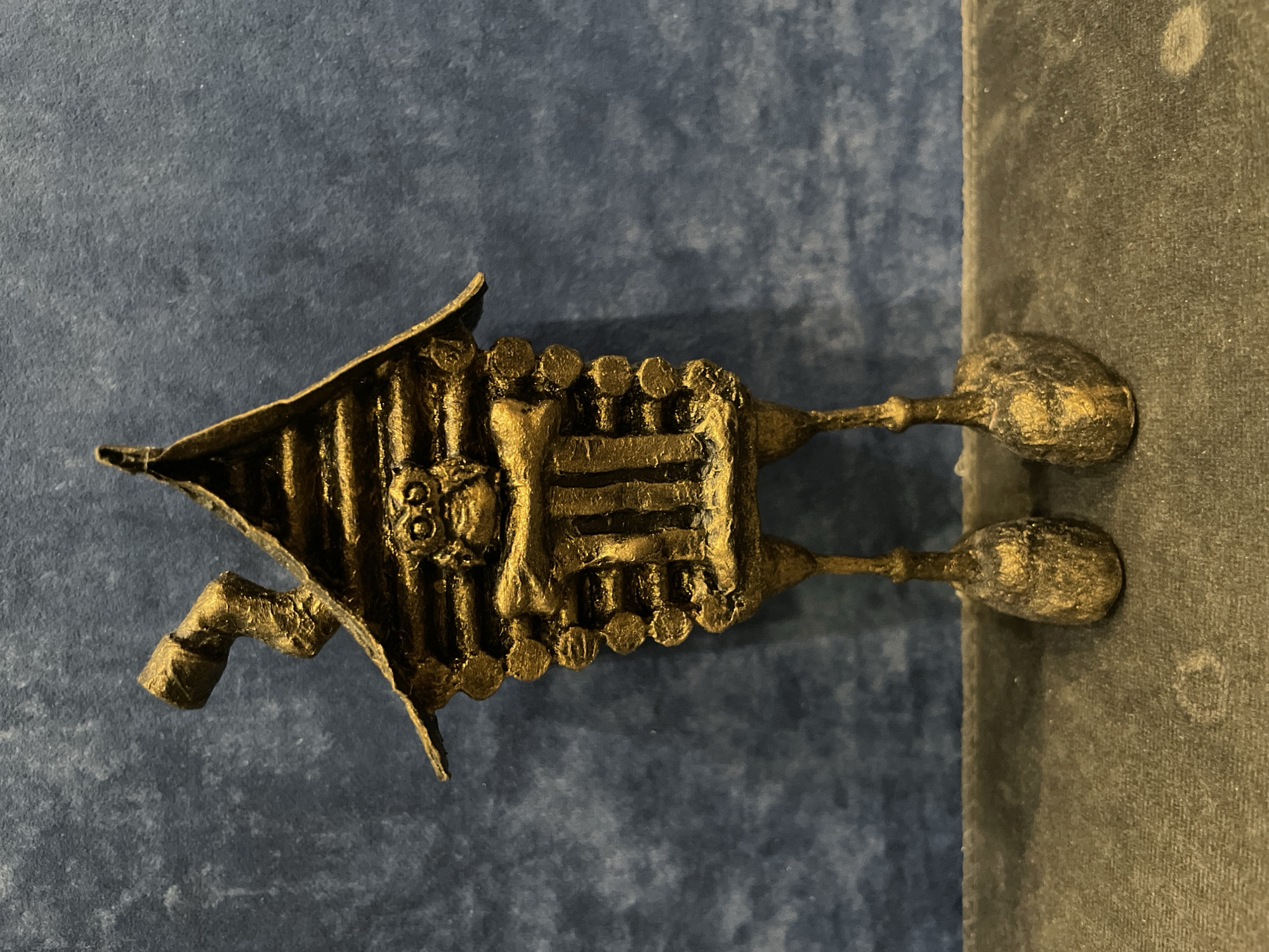 A gold-colored, whimsical figurine of a log cabin with a chimney and legs stands against a textured blue background. The figurine has a face with large, round eyes and a mouth carved into the "log" front. The cabin is made to resemble stacked logs, with a peaked roof. The legs are thin and spindly, ending in bulbous feet. The figurine appears to be made of a solid material, possibly metal or painted plaster. It is positioned on a dark, textured surface. The setting does not suggest a specific location in the city of Perm, Russia.