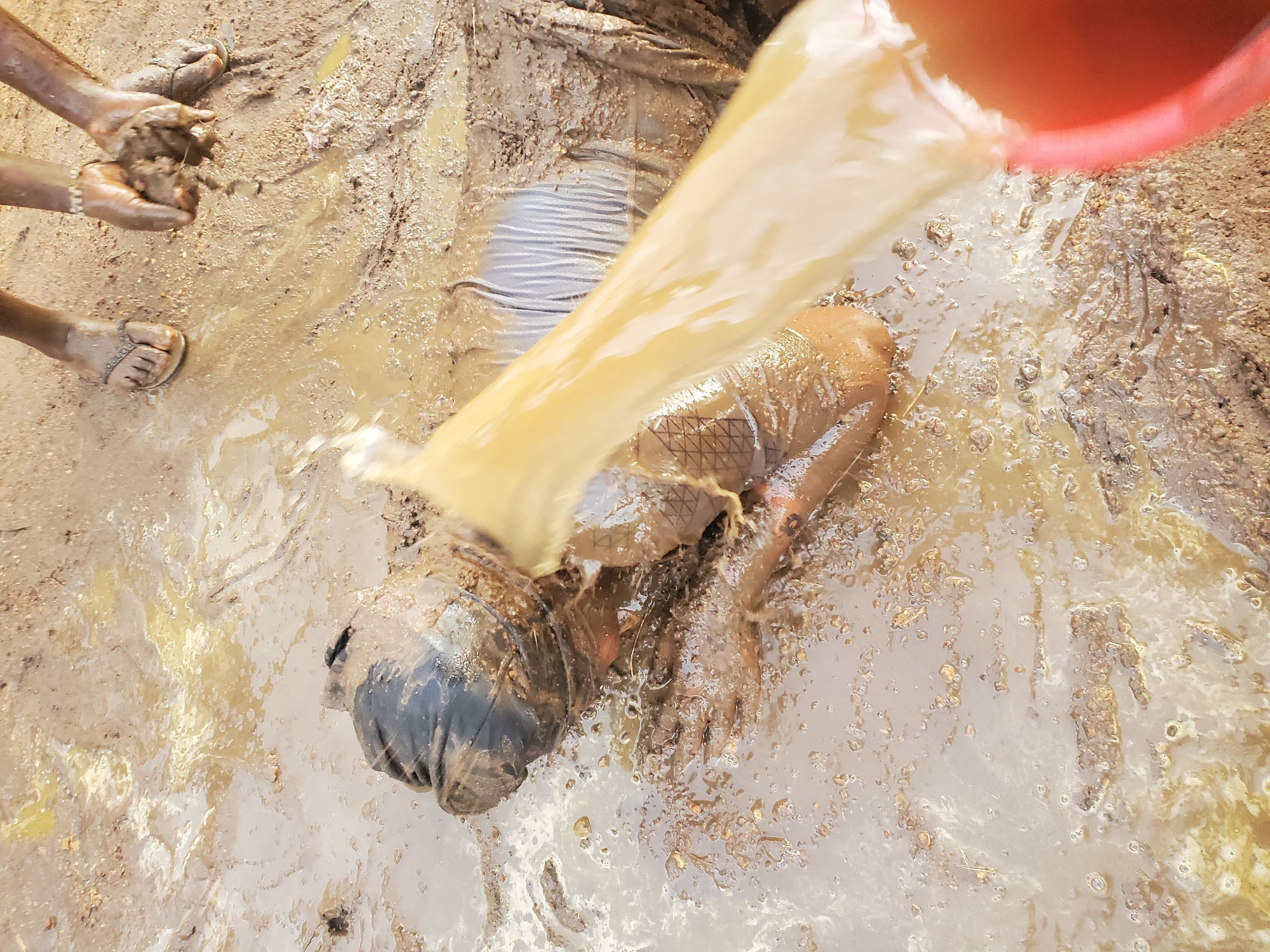 This high-angle shot captures a dynamic scene on muddy ground in Akwanga, Nigeria. The primary subject is a person, likely a child, lying face down and largely submerged in thick, brown mud. This individual is dressed in dark-colored clothing, with light-colored undergarments or a pattern visible on their lower back.
From the upper right, a stream of murky, yellowish-brown liquid pours from a red plastic bucket, dousing the head and upper body of the person in the mud. To the left, another person's hands are visible, covered in mud and actively gathering or playing with clumps of it. Their feet, clad in light-colored sandals, are also visible, standing in the muddy mixture.
The ground is saturated and uneven, composed of wet soil, mud, and small pebbles, creating a very messy environment. The overall impression is one of an outdoor activity, possibly a form of play or a customary washing in a muddy area. The lighting suggests it is daytime. There is no visible text in the image.