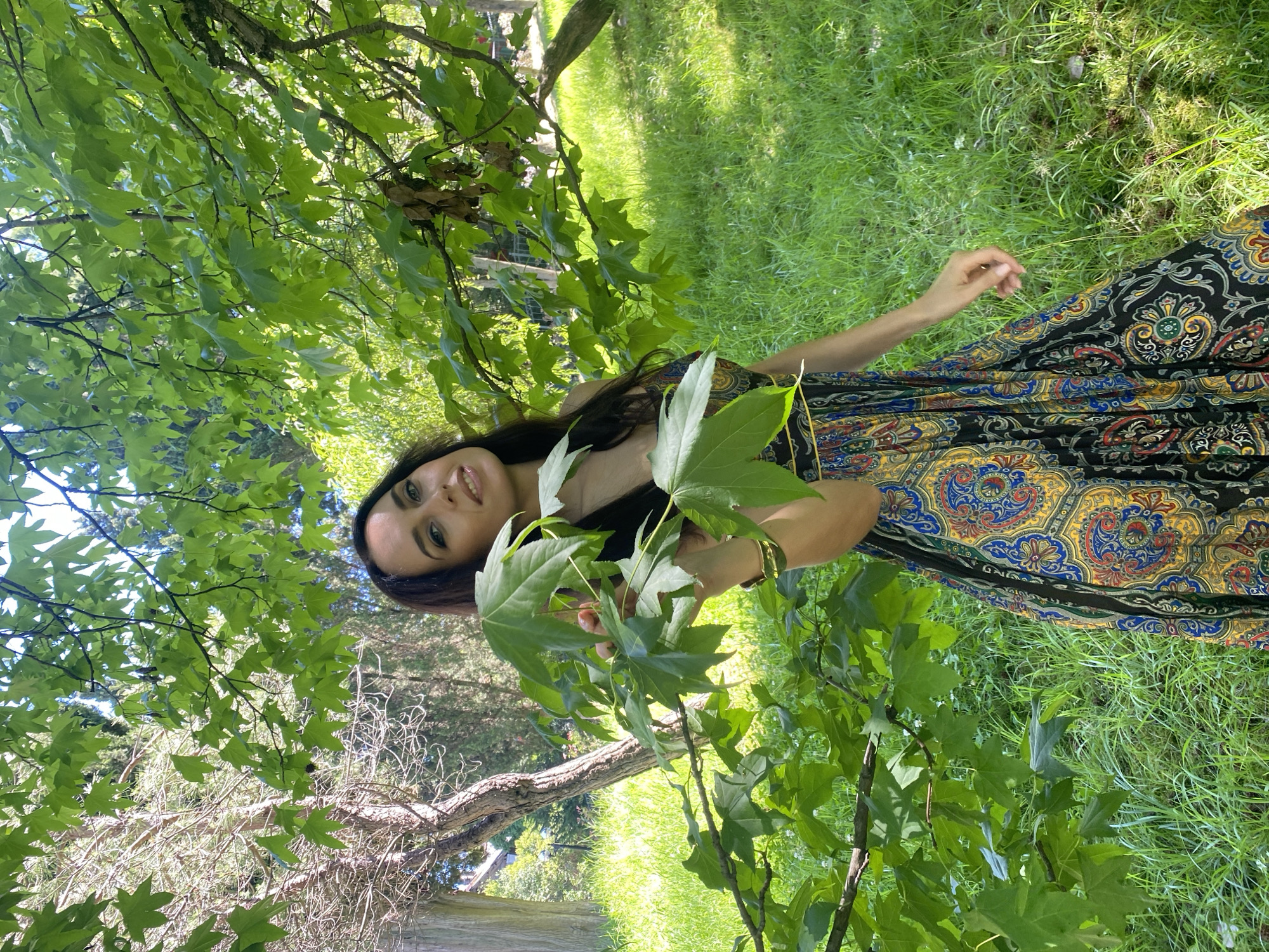 A young woman with dark hair and a warm smile is pictured in a lush, green outdoor setting. She is wearing a long, flowing dress with a vibrant paisley pattern in shades of blue, yellow, and red. Her right arm is extended, and she is gently holding several large, green leaves.
The scene appears to be a park or a garden, with abundant green grass and the dappled sunlight filtering through the leaves of trees. The lighting suggests it is daytime, likely late morning or early afternoon, and the weather seems pleasant. The woman’s relaxed posture and happy expression convey a sense of peace and enjoyment of nature. The image is shot from a low angle, emphasizing the surrounding foliage and the woman's presence within it. No visible text is present in the image. The location context provided is Sochi, Russia.