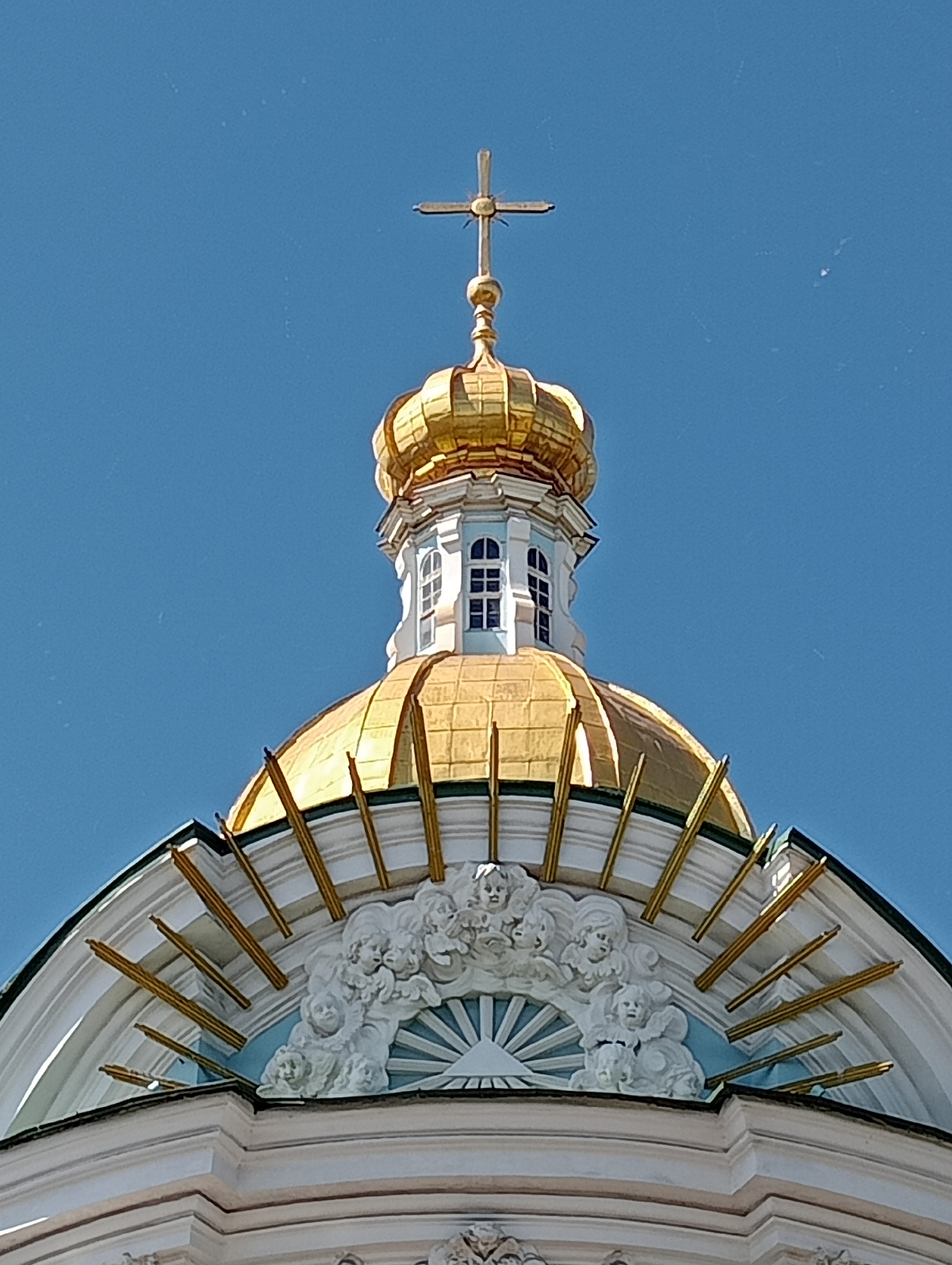 The image shows the golden dome and cross of a church, likely St. Nicholas Naval Cathedral in St. Petersburg, Russia. The dome is ornate, with decorative elements around its base. A bright blue sky serves as the background, with a few faint dust specks visible. The architectural style suggests a religious building.