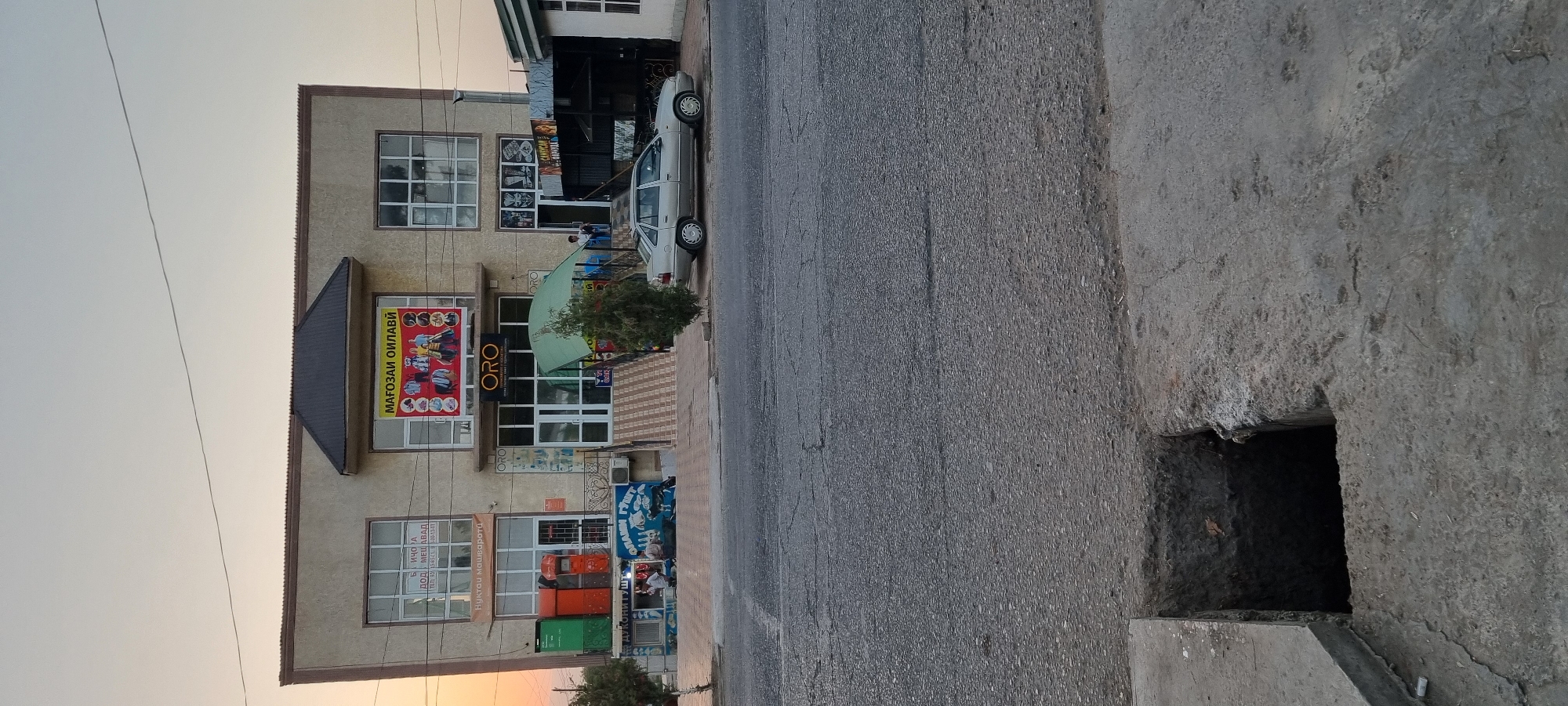 In the city of Ismoili Somoni, Tajikistan, a two-story building with a beige facade stands prominently. The upper facade features large windows, and a prominent sign with text "МАГОЗАИ ОИЛАВИ" and images of clothing is displayed above the entrance. Below this, a sign reads "ORO" in large letters. The ground floor has multiple storefronts. To the left, there are three bright-colored mailboxes (green, orange, and red) next to a shop. To the right of the main entrance, a green awning covers a set of stairs leading up to the building. A silver sedan is parked in front of the building. The street in front of the building is paved and shows signs of wear. A square hole has been dug into the concrete sidewalk in the foreground. The sky above is a pale orange, suggesting it is either sunrise or sunset. A few trees are visible near the building.