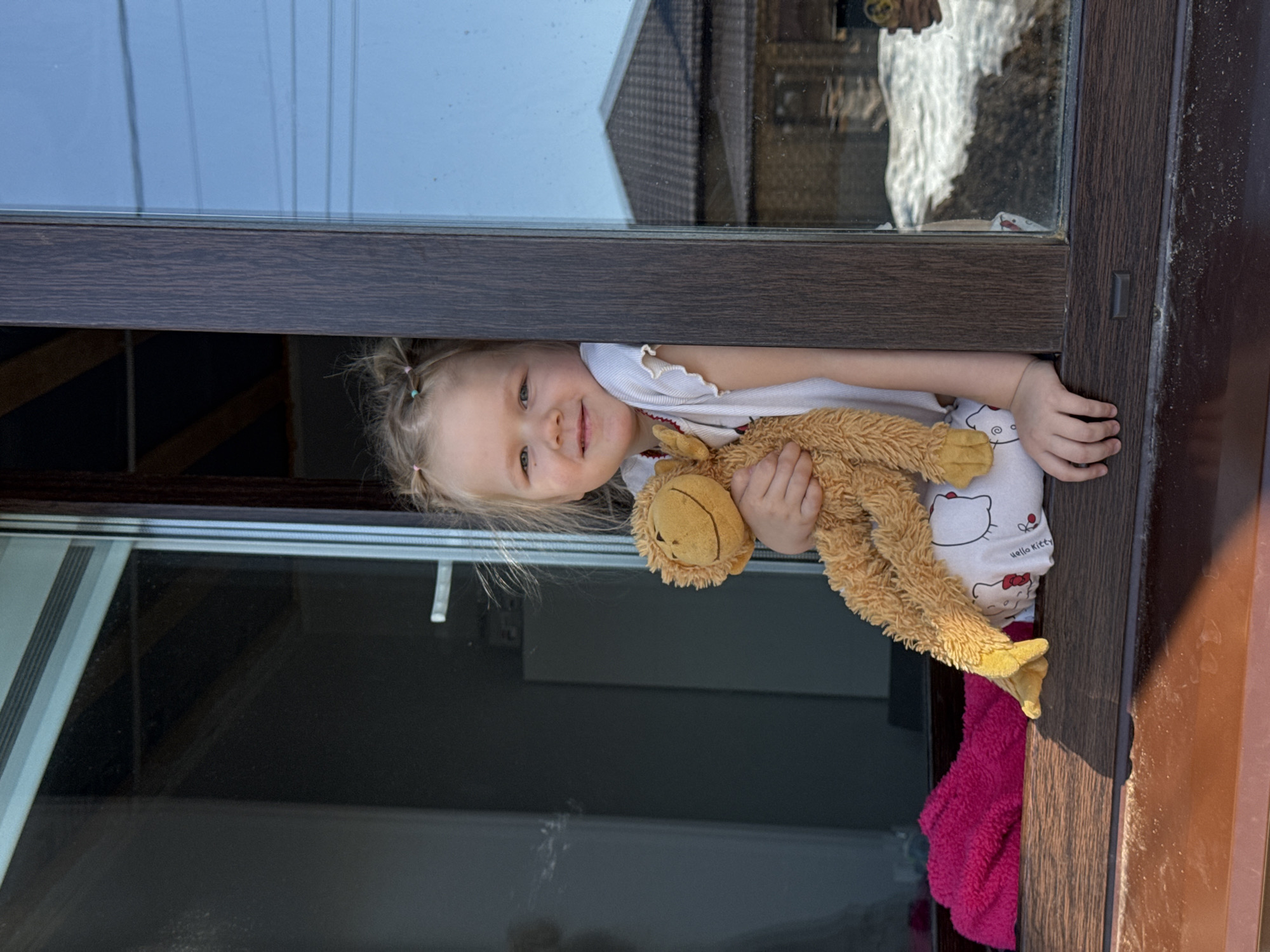 A young girl with blonde hair in pigtails is looking out a window in Gornyi (Dvurechenskoe S/P), Russia. She is holding a stuffed monkey. She is wearing white Hello Kitty pajamas. A red blanket is next to her at the bottom of the window. The window has a dark brown frame. Outside of the window are buildings with reflective glass.