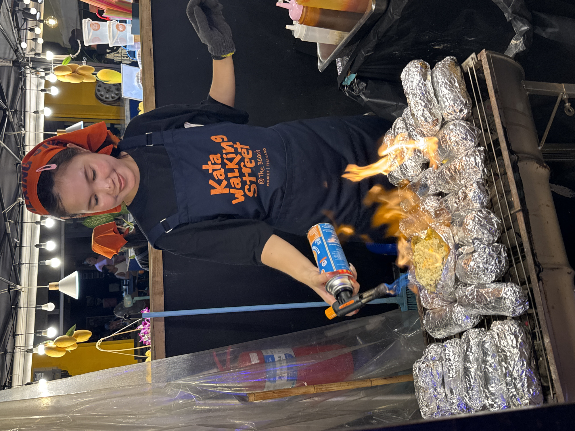A young woman with a bright orange bandana and a dark blue apron is the central subject of this image. She is smiling and actively using a blowtorch, its flame directed at a rack of food items wrapped in aluminum foil. The food items appear to be cylindrical, possibly corn or potatoes, arranged on a metal grill. The woman wears a grey glove on her left hand, holding the blowtorch with her right. The setting appears to be an outdoor food stall at night, with strings of lights visible in the background. There are also what appear to be bottles of sauces or condiments on a tray behind her, and a trash bag. The apron has visible text that reads: "Kata Walking Street @ The Beach Phuket, Thailand". The overall atmosphere suggests a lively street food scene in Karon, Thailand. The woman's expression conveys enthusiasm and engagement with her task.